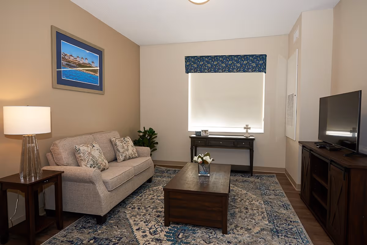 A cozy living room with a beige sofa adorned with patterned cushions, a wooden coffee table with a small flower arrangement, a side table with a lamp, a framed picture on the wall, a window with a floral valance and a roller shade, a dark wooden console table beneath the window, and a flat-screen TV on a wooden TV stand. The room features a patterned area rug and neutral-colored walls.