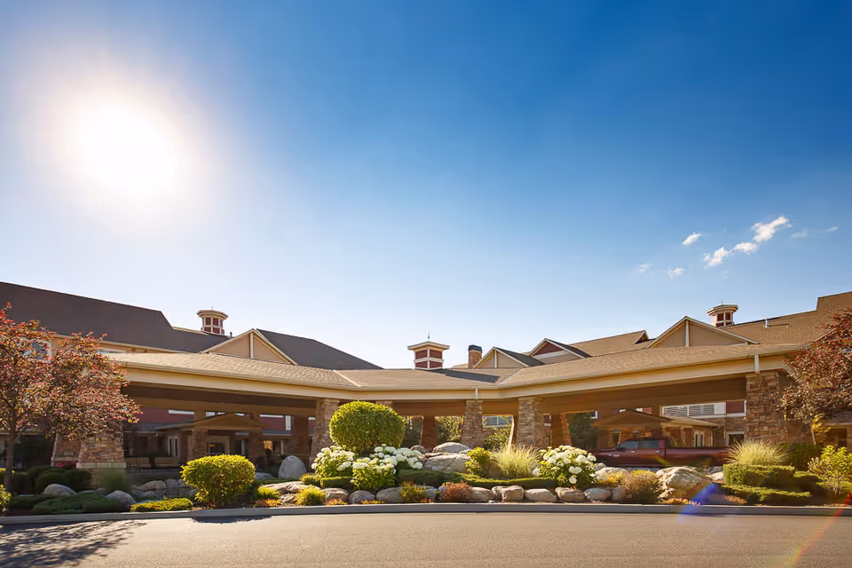 Exterior view of The Bridge At Post Falls senior living facility on a sunny day, showing a covered entrance with stone pillars, landscaped bushes, flowers, and a clear blue sky.