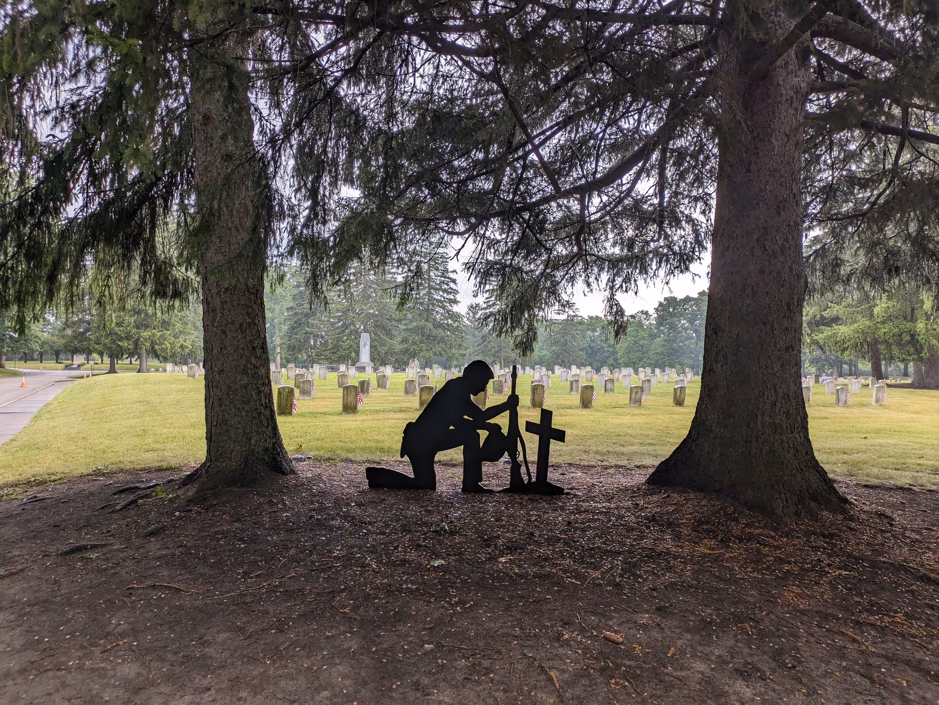 Silhouette of a person kneeling by a cross in a cemetery beneath two large trees