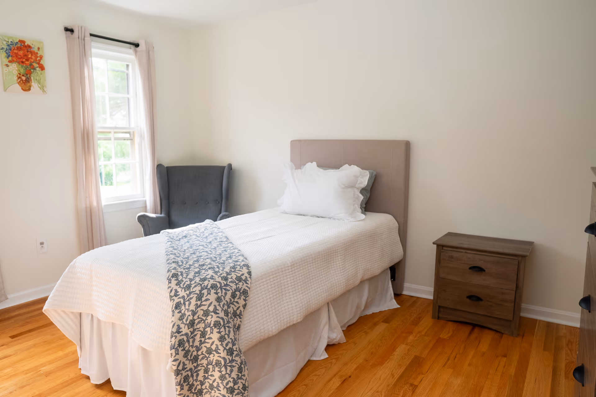 A tidy bedroom with a bed, chair, nightstand, and window letting in natural light.