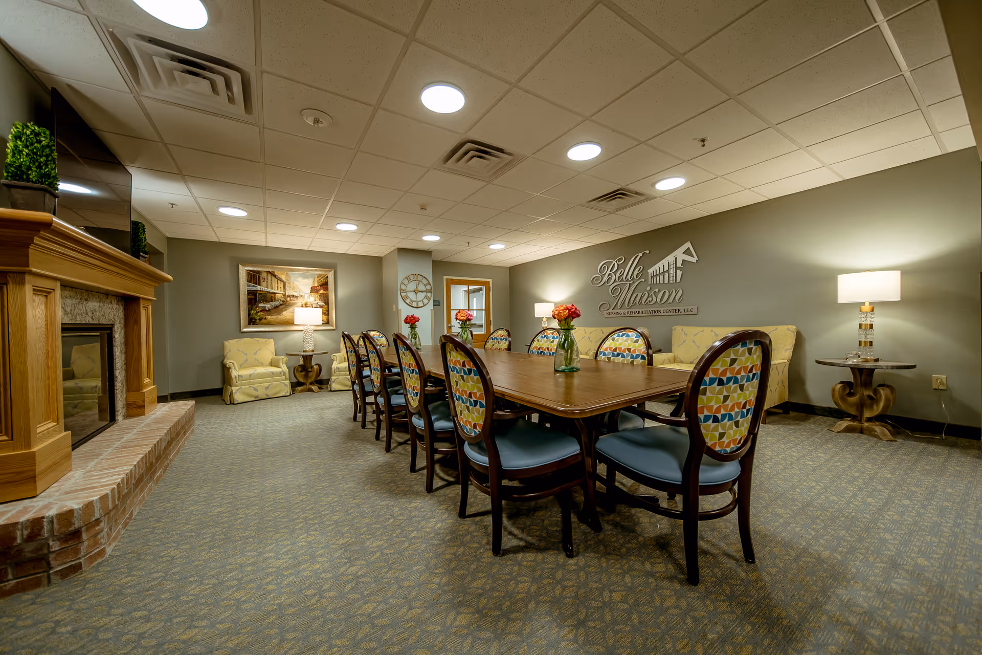 A cozy common area with a long wooden table surrounded by chairs with colorful patterned backs. The room features a fireplace with a wooden mantle on the left, two armchairs with a side table and lamp in the back corner, and a wall with the Belle Maison Nursing & Rehabilitation Center, LLC logo. The carpeted floor and ceiling lights create a warm and inviting atmosphere.