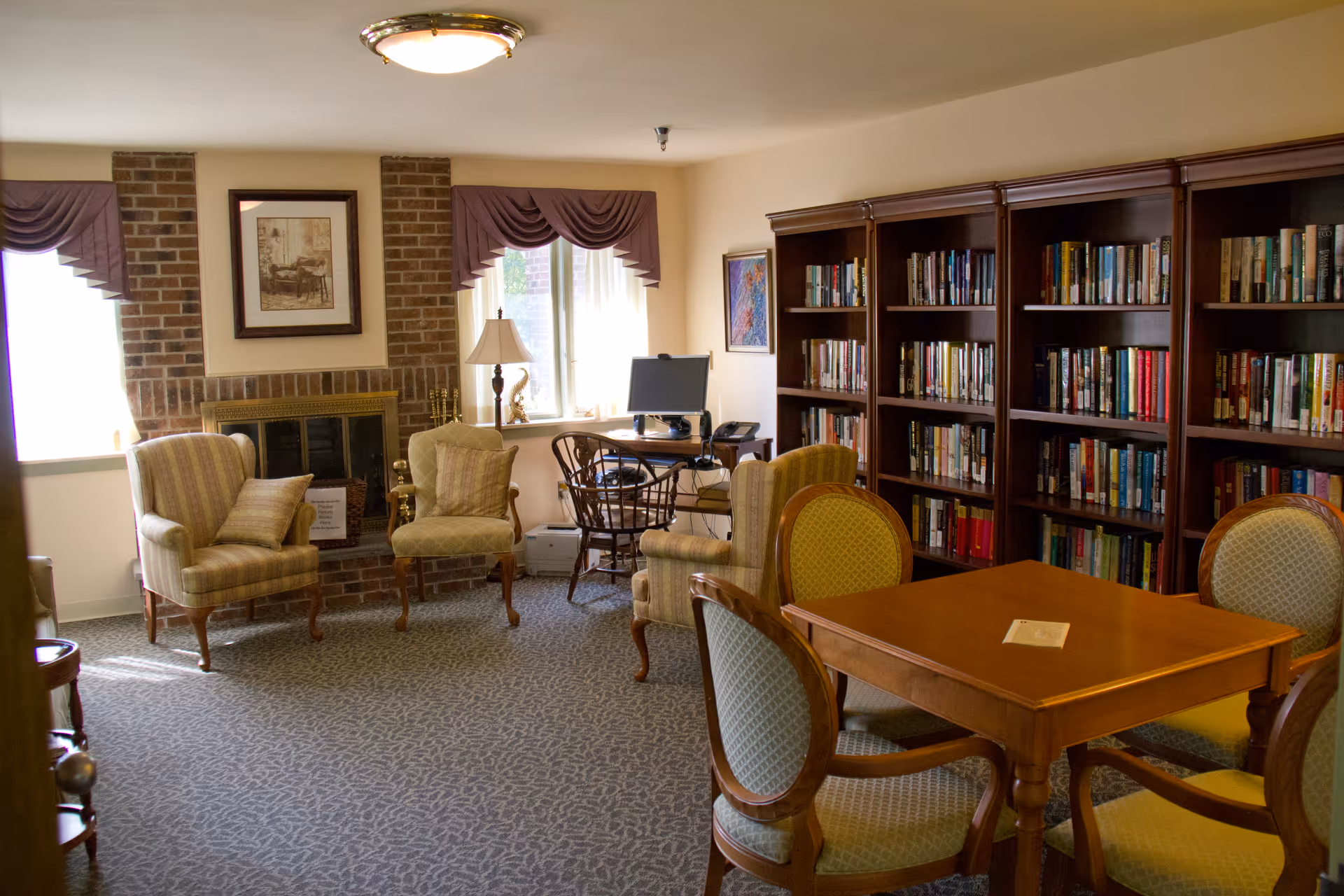 A cozy library room with a brick fireplace, two armchairs with cushions, a wooden table surrounded by four upholstered chairs, a computer desk with a chair, and large bookshelves filled with books. The room has carpeted flooring and windows with purple valances allowing natural light to enter.