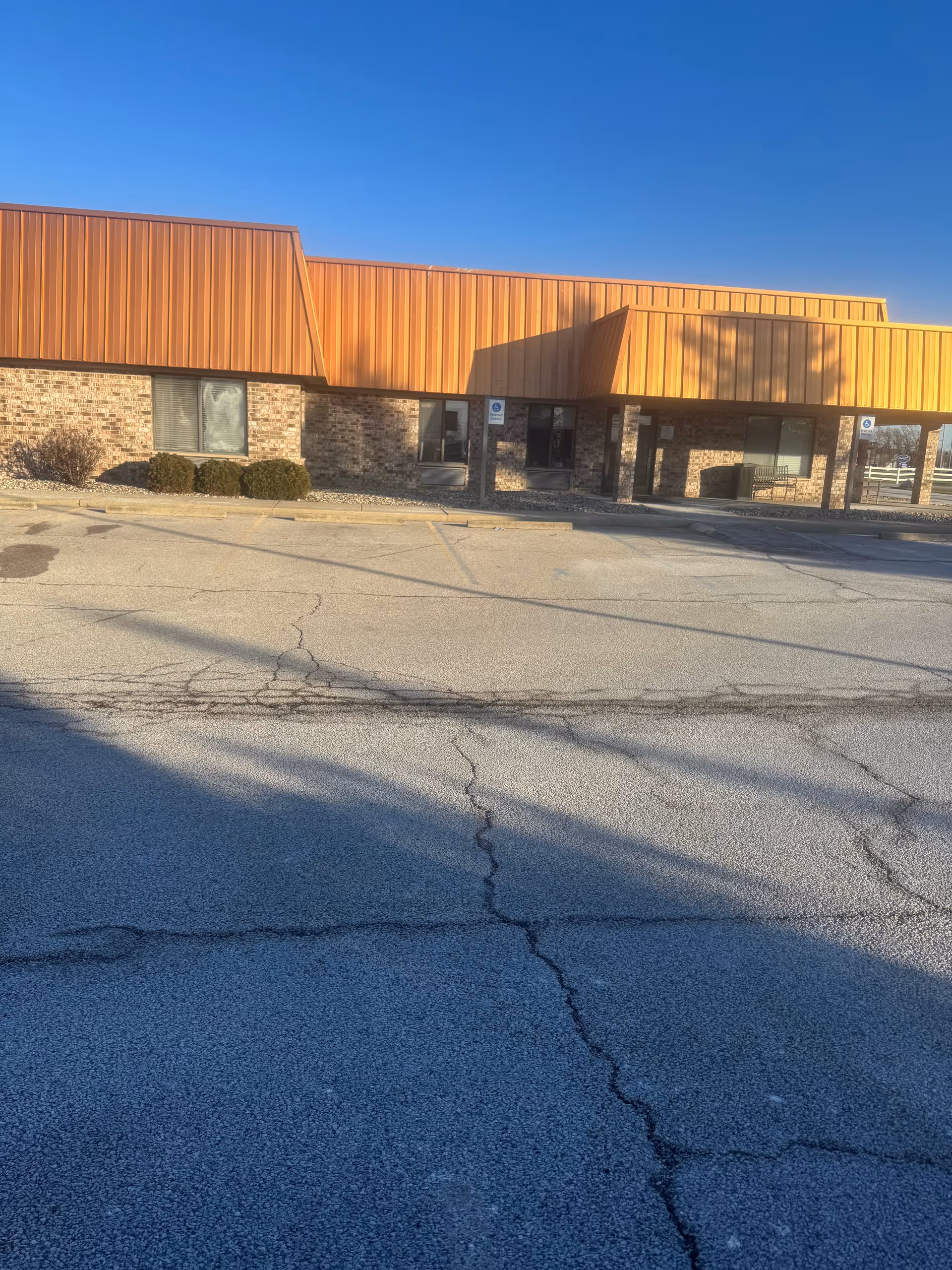 Front exterior of a brick facility with orange metal roofing and a parking lot.