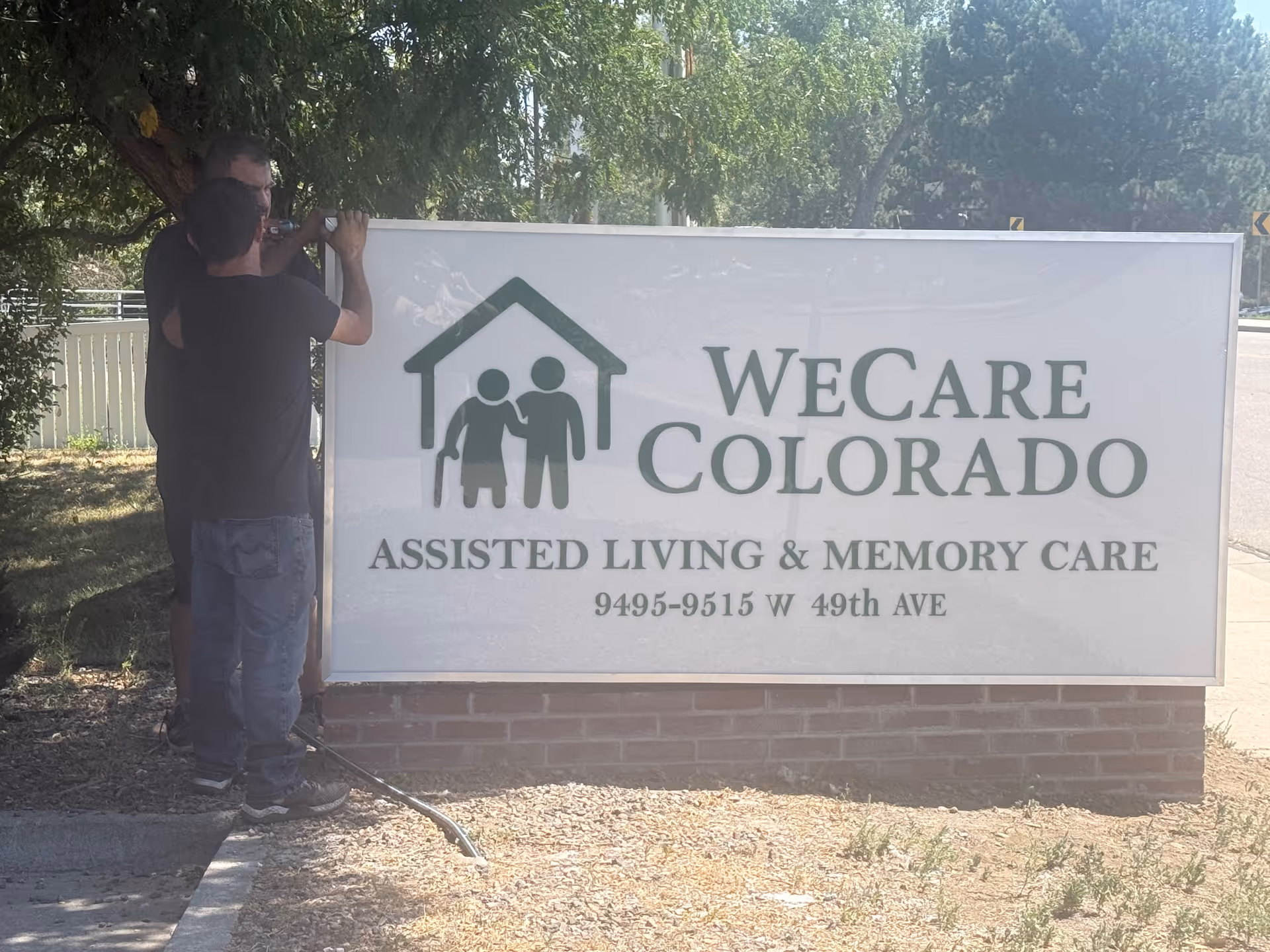Two men adjust a large outdoor sign reading WeCare Colorado Assisted Living & Memory Care.