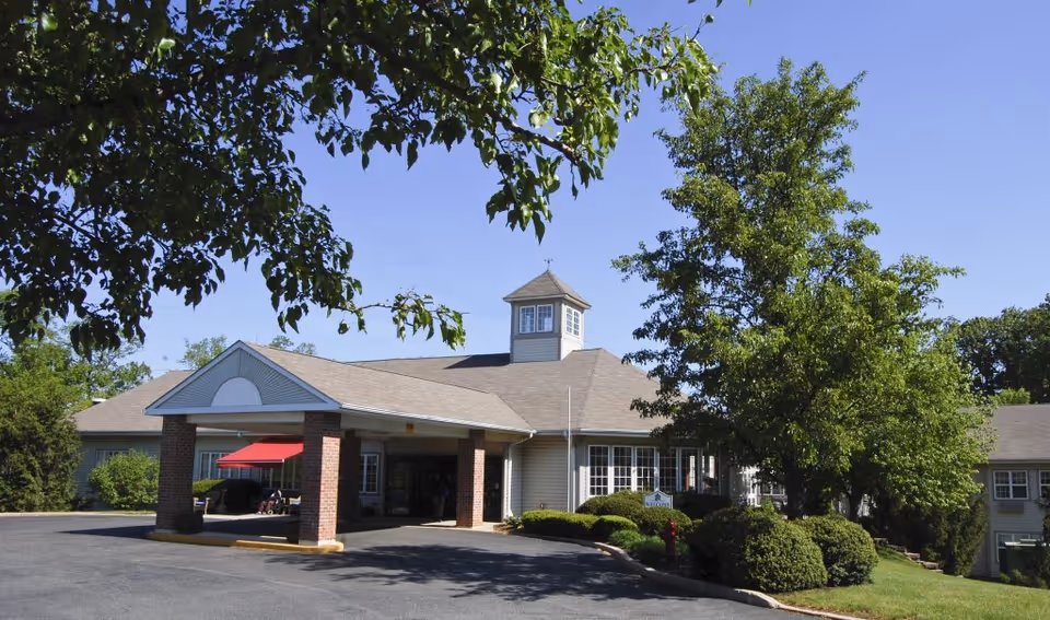 Front entrance of a single-story assisted living building with a covered porte-cochère, a red awning, and trees and landscaping.
