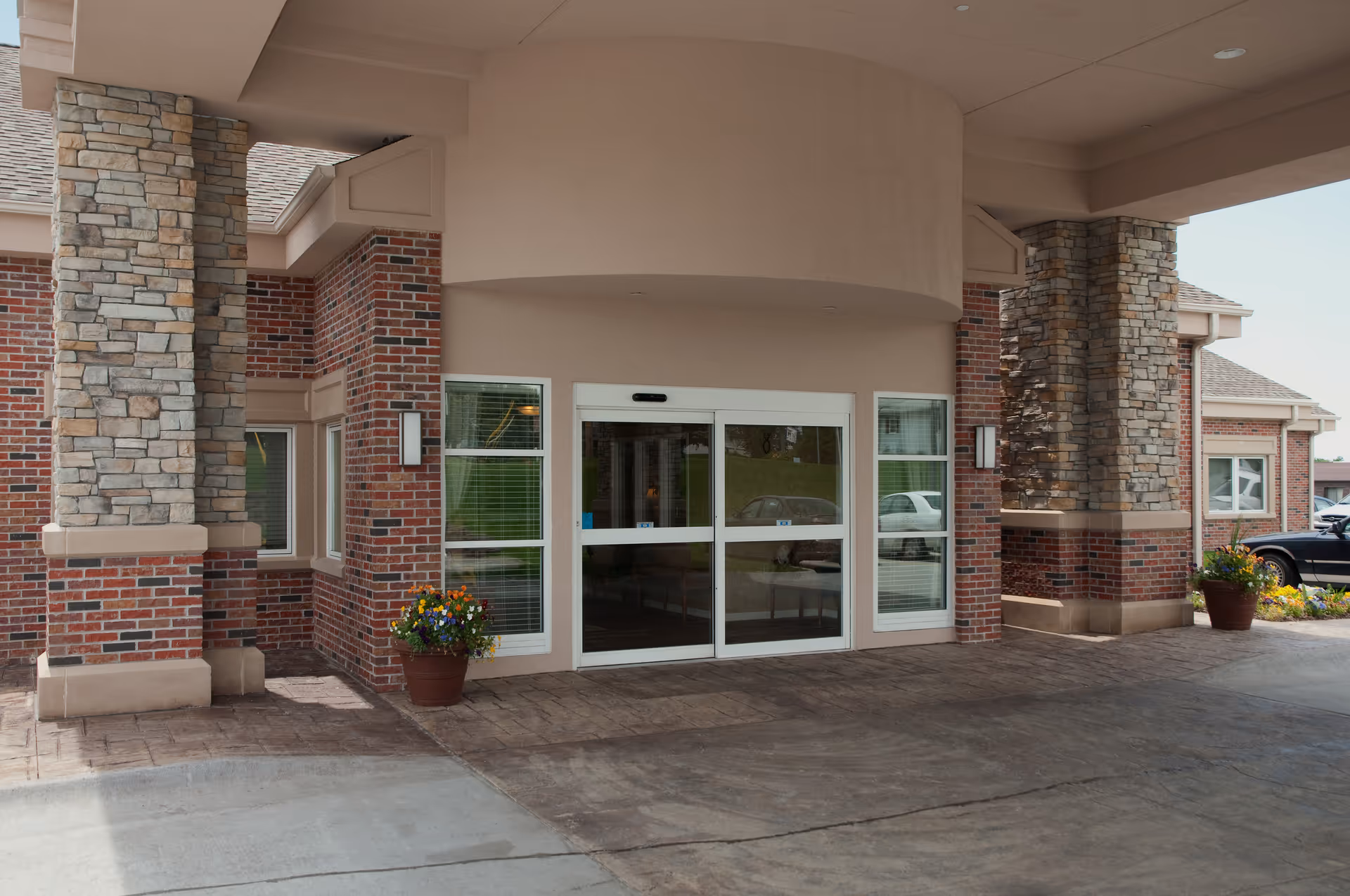 Covered entrance of a brick-and-stone senior living facility with sliding glass doors and potted flower pots.