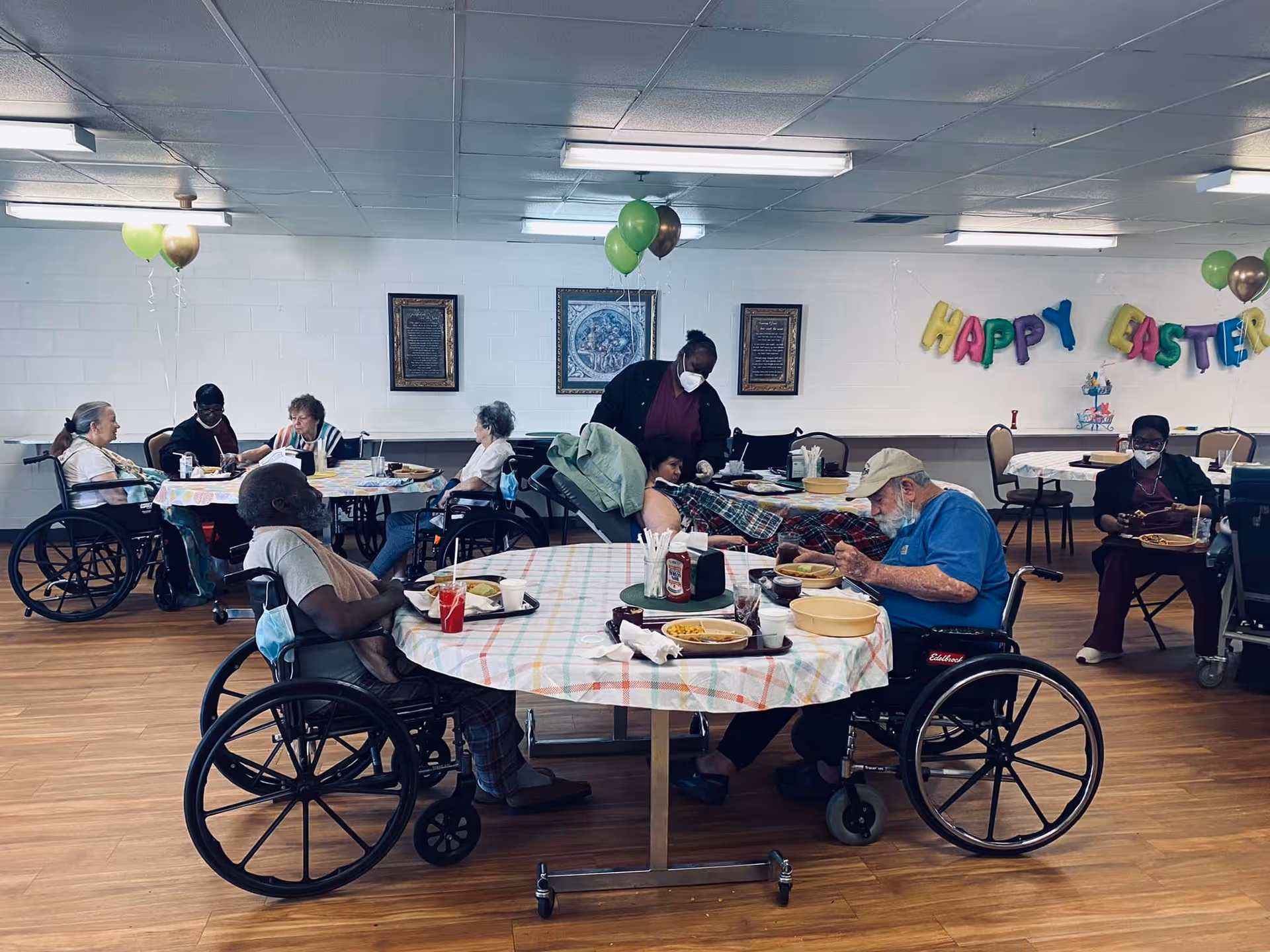 Elderly residents in wheelchairs sit around decorated tables eating and socializing in a nursing home dining room with a 'Happy Easter' banner.