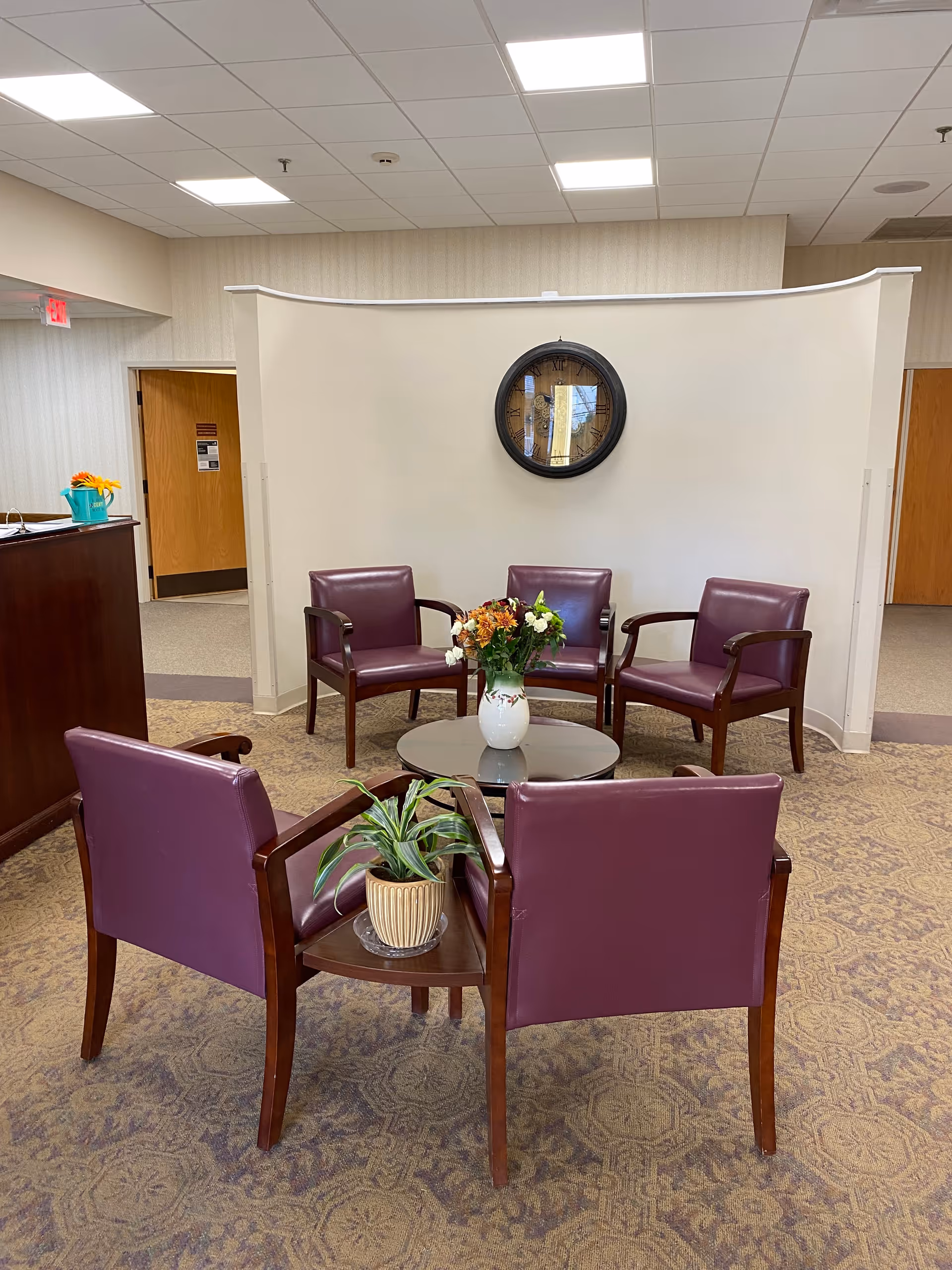 A seating area in a senior living facility with five purple armchairs arranged around a small round table with a vase of flowers. There is a potted plant on a small side table attached to one of the chairs. A large round clock hangs on a curved beige wall behind the chairs. The room has patterned carpet and a drop ceiling with fluorescent lights.
