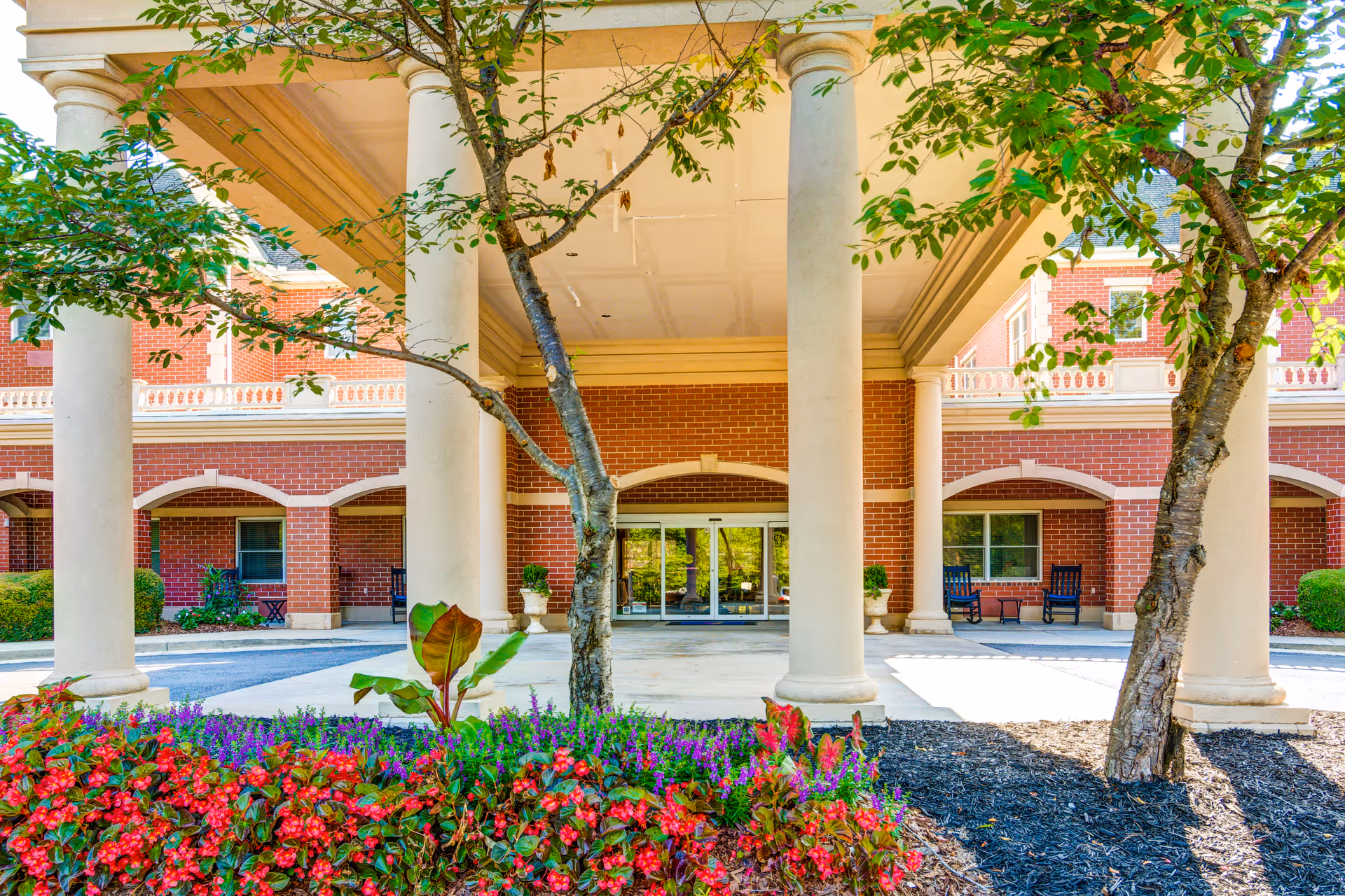 Entrance area of Dunwoody Place featuring large white columns supporting a covered driveway. There are two trees and colorful flower beds with red and purple flowers in the foreground. The building has a red brick exterior with white trim and several windows. Two black rocking chairs are visible on the porch area near the entrance.