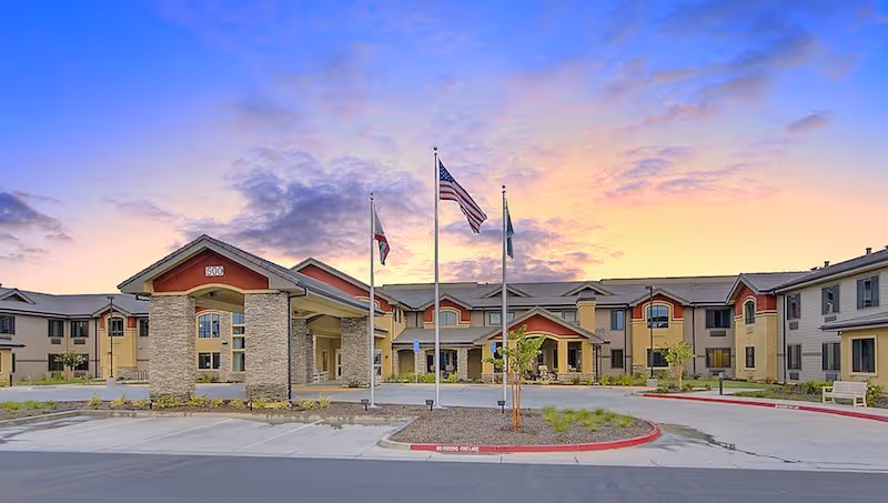 Front view of The Pines, A Merrill Gardens Community, with flags and a colorful morning sky.