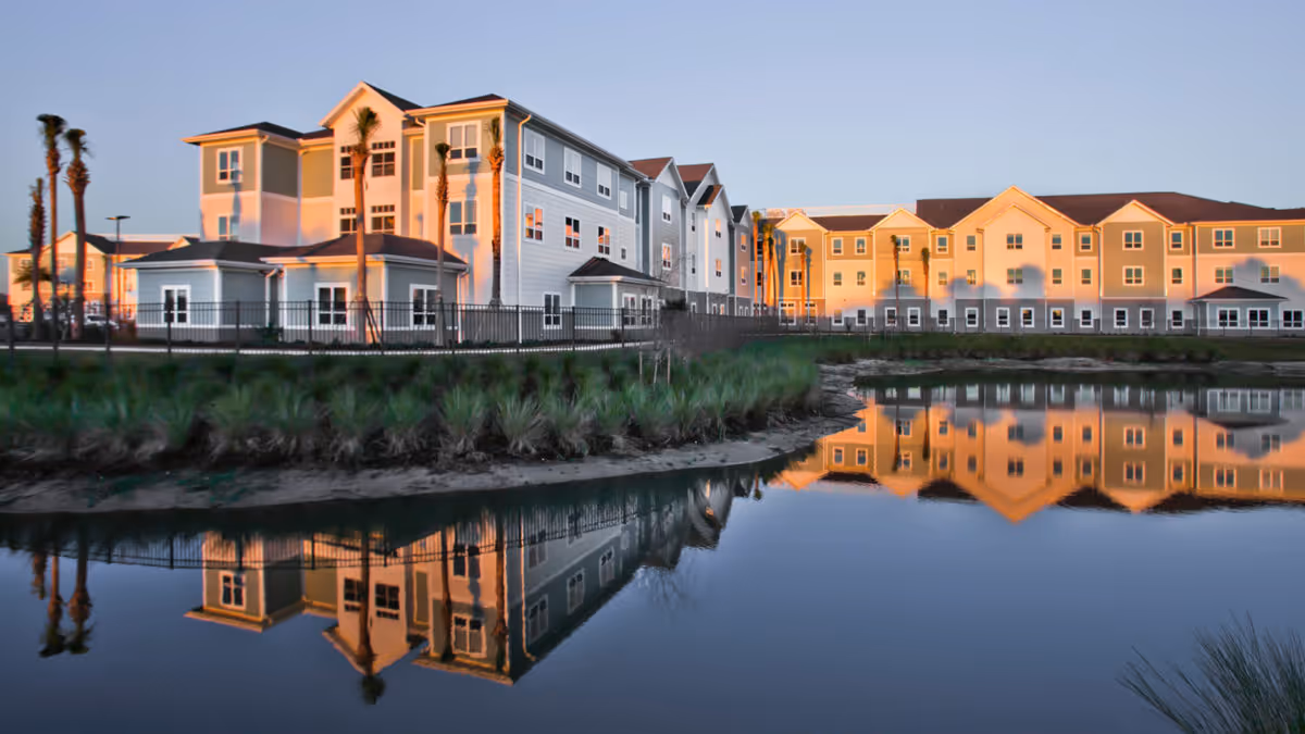 Three-story senior living building by a pond with its reflection in the water at sunset.