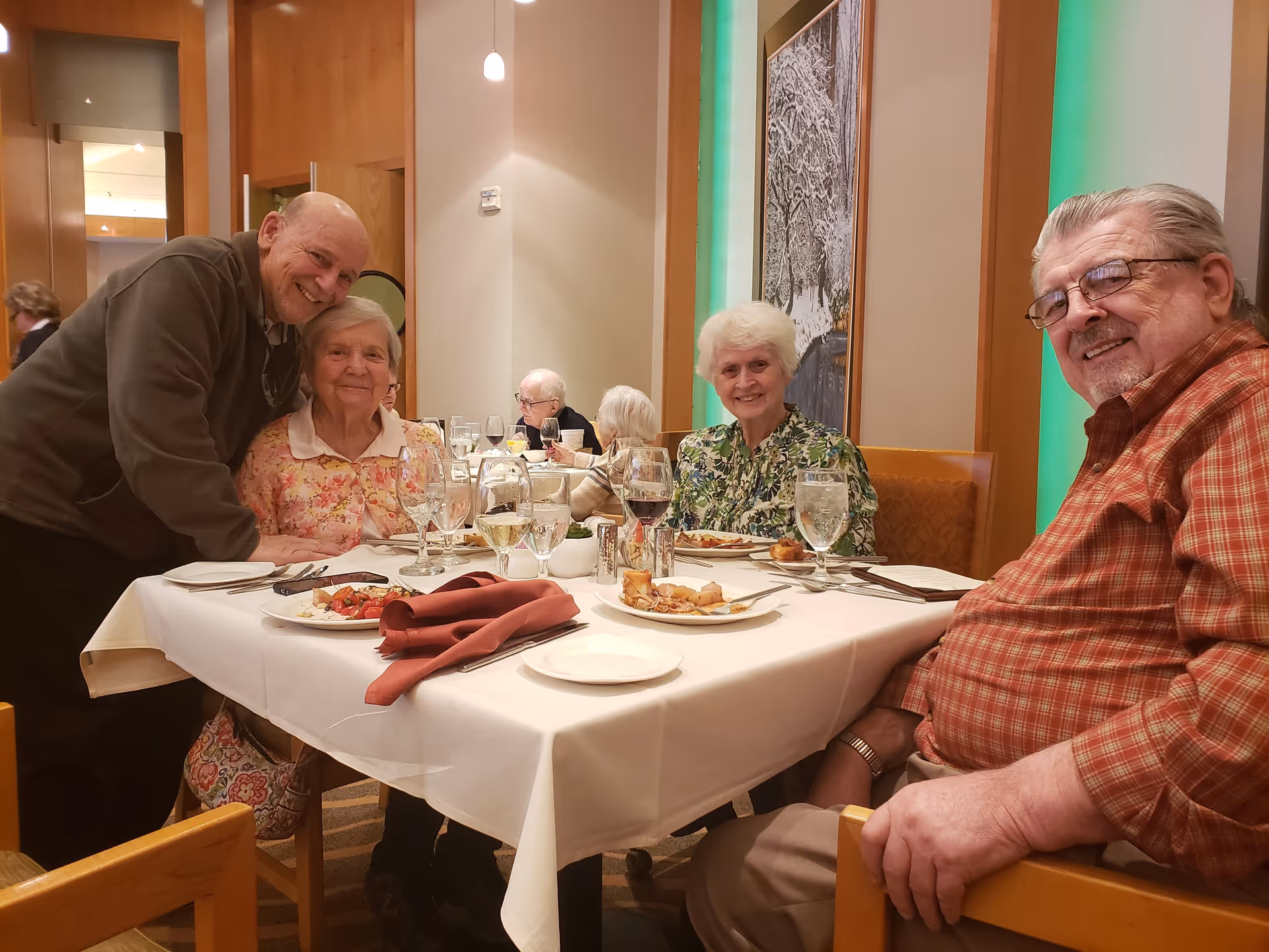 Four elderly people sitting around a dining table in a well-lit room, smiling at the camera. The table is set with plates of food, glasses of water and wine, and napkins. The background shows a wall with a large framed picture and green accent lighting.
