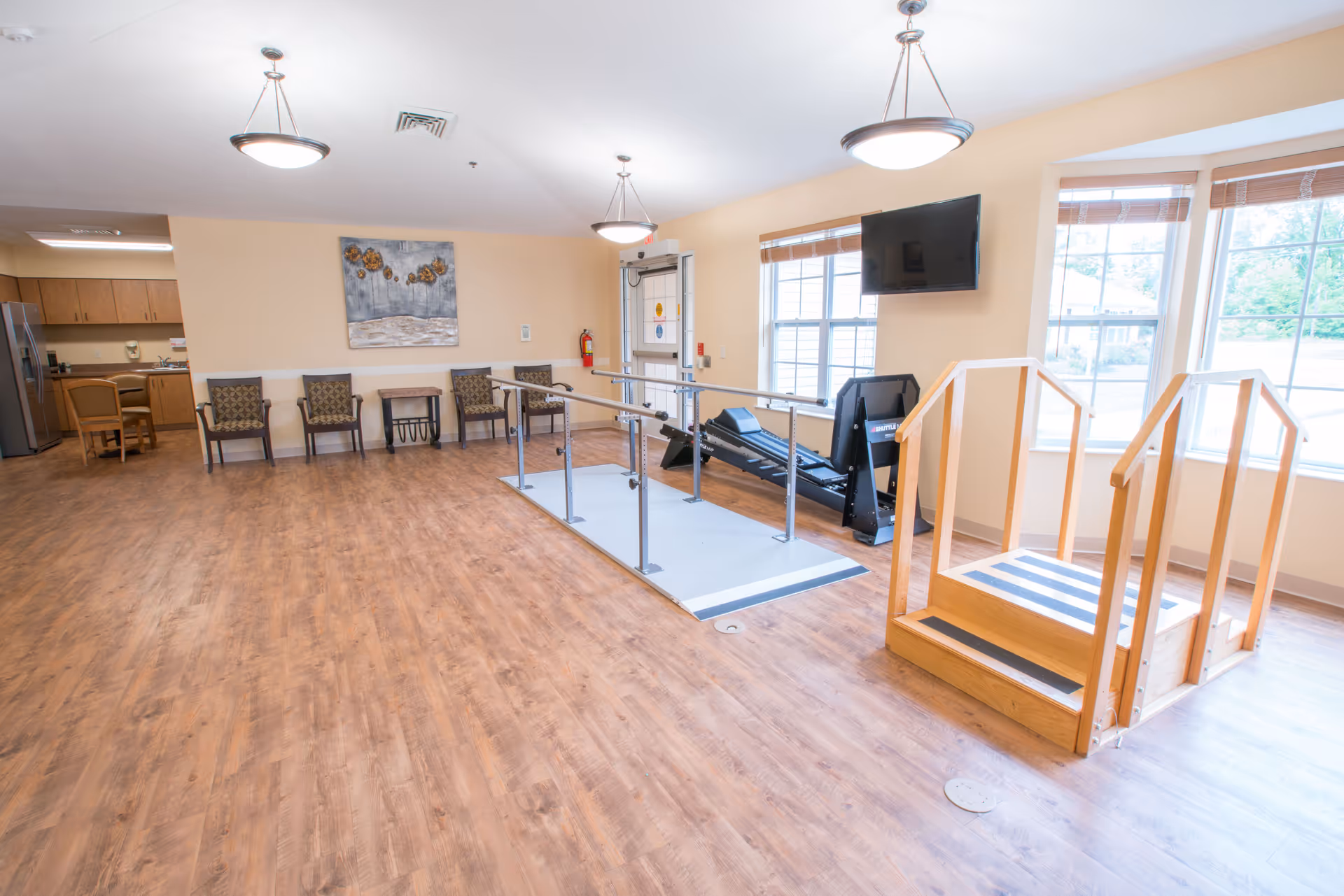 A spacious room with wooden flooring featuring a set of parallel bars and a wooden staircase for physical therapy or rehabilitation. There are several chairs lined up against a beige wall with a decorative painting above them. A flat-screen TV is mounted on the wall near large windows letting in natural light. In the background, there is a kitchen area with wooden cabinets and a refrigerator.