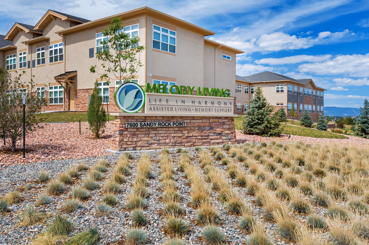 Exterior view of Melody Living senior living facility with a large sign in front displaying the name, slogan 'Life in Harmony', and services 'Assisted Living, Memory Support'. The building is beige with multiple windows and surrounded by landscaped grounds with small trees and shrubs under a partly cloudy blue sky.