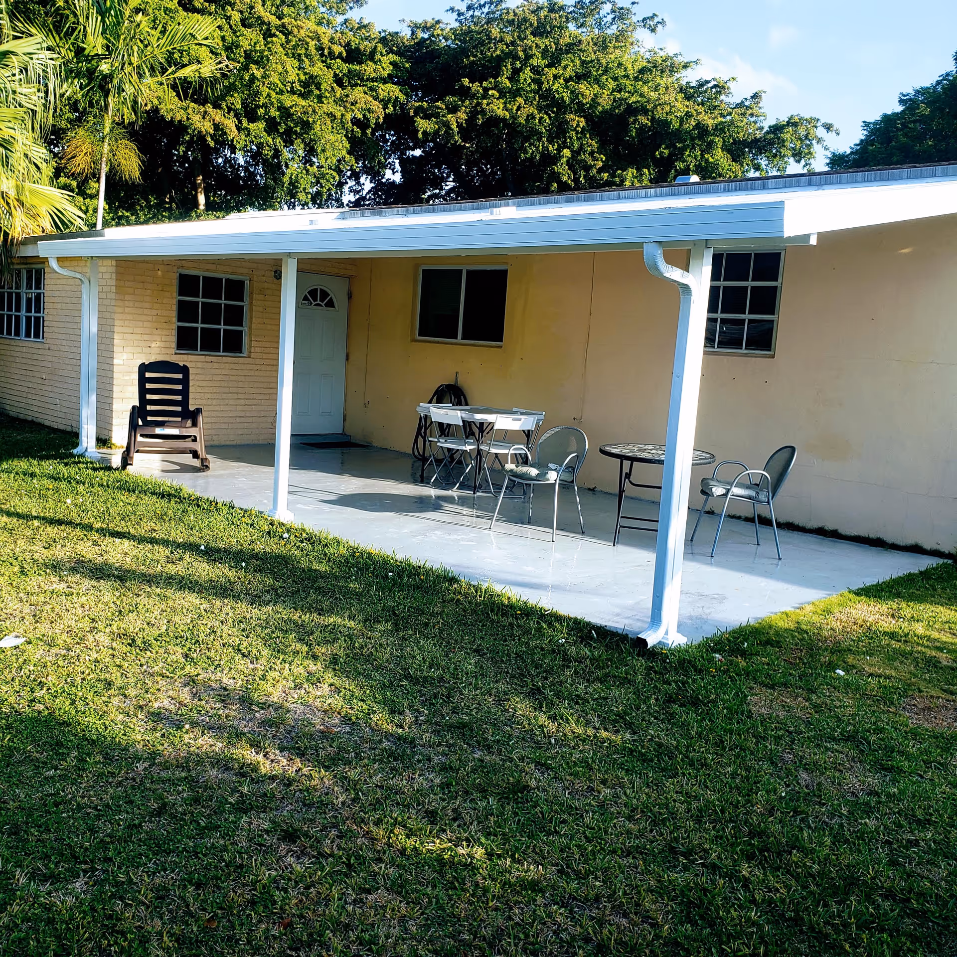 Outdoor patio area of a building with a covered roof supported by white posts. There are two sets of tables and chairs on the concrete patio, surrounded by green grass and trees in the background.