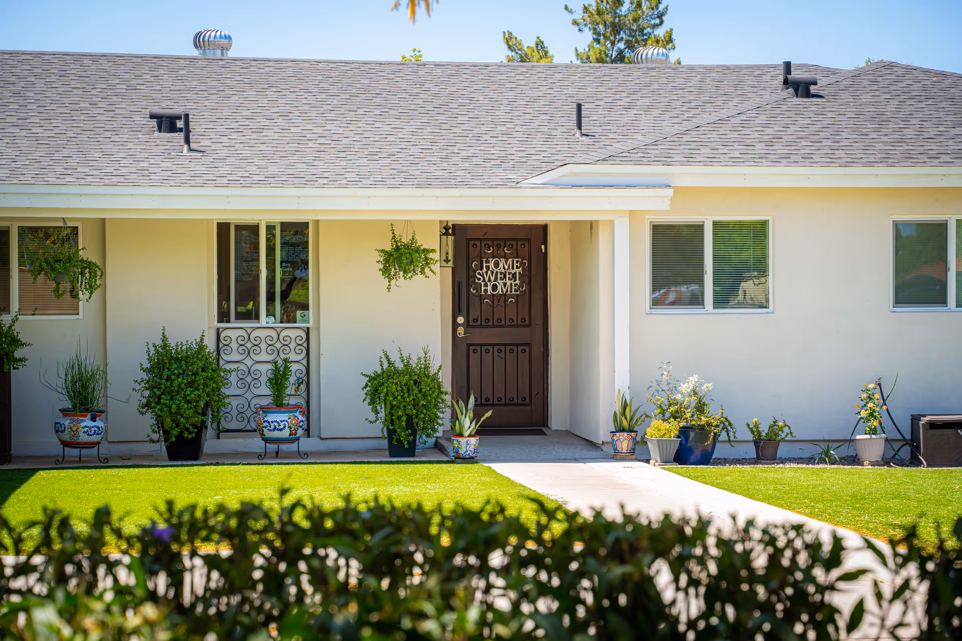 Front exterior view of a single-story house with a brown door that has a 'Home Sweet Home' sign. The house has beige walls, a gray shingled roof, several windows, and a variety of potted plants and hanging plants near the entrance. There is a well-maintained green lawn and a concrete pathway leading to the door.