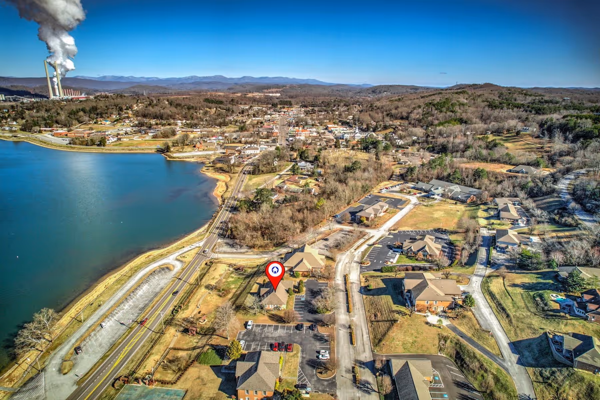 Aerial view of a small town with a lake on the left side and multiple buildings spread out along roads. There is a red location pin marking a specific building near the lake. In the background, there are hills and a factory with large smokestacks emitting white smoke.