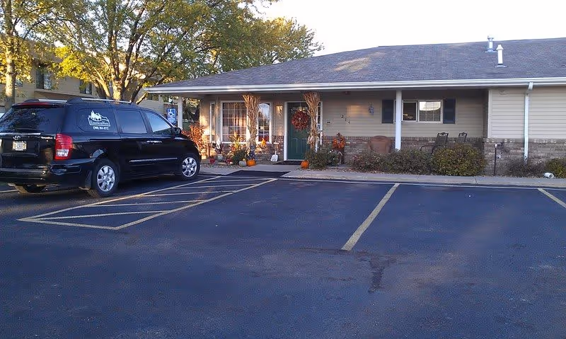 Exterior view of a single-story building with a green door decorated with a fall wreath and surrounded by autumn-themed decorations including pumpkins and cornstalks. A black vehicle is parked in front of the building in a parking lot with marked spaces. Trees with green and yellow leaves are visible in the background.