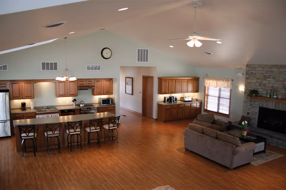 Open communal interior with a long kitchen island and bar stools, a seating area, and a stone fireplace.