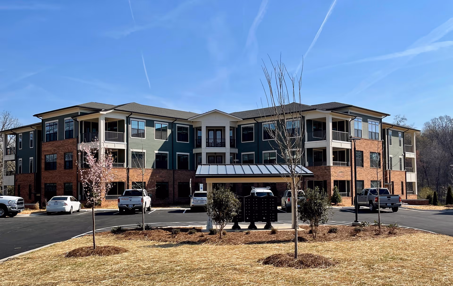 Exterior view of a three-story senior living facility building with green and brick facade, multiple windows, balconies, and a covered entrance. There are several parked cars in front, young trees, and a clear blue sky above.