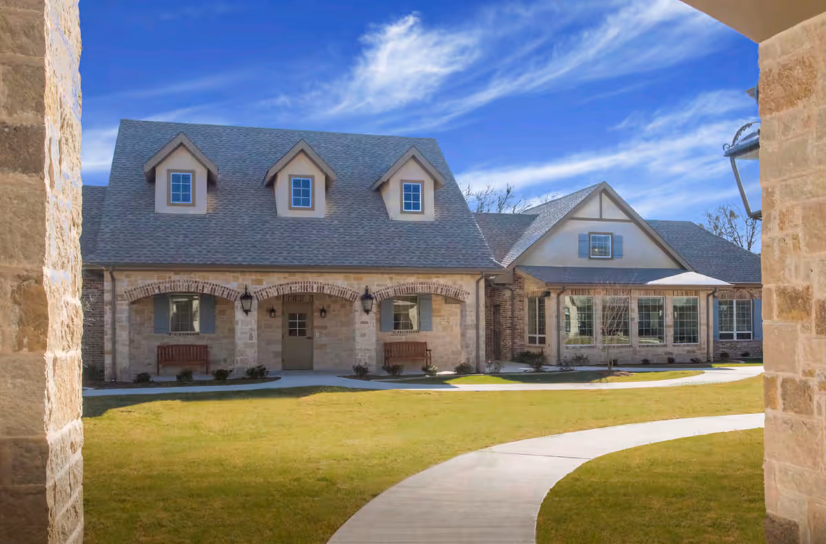 Stone-front cottage building with dormer windows, arched porch and benches viewed across a grassy courtyard with a curved walkway.