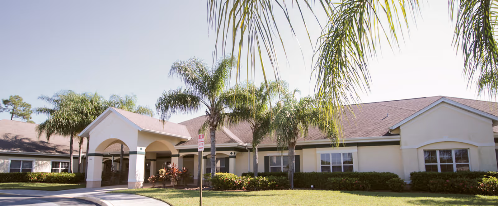 Exterior view of a single-story senior living facility building with a covered entrance, surrounded by palm trees and well-maintained landscaping under a clear sky.