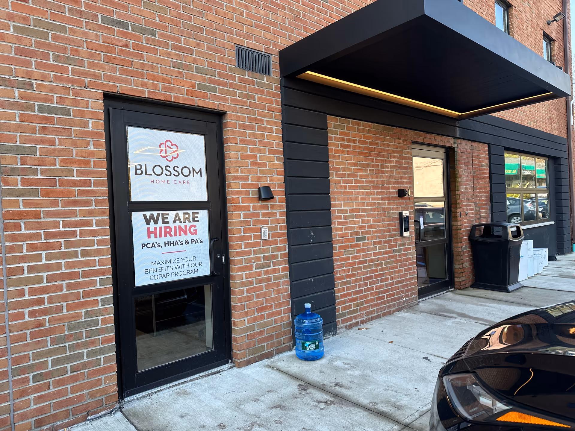 Brick building entrance of Blossom Home Care with a hiring sign and blue water jug.