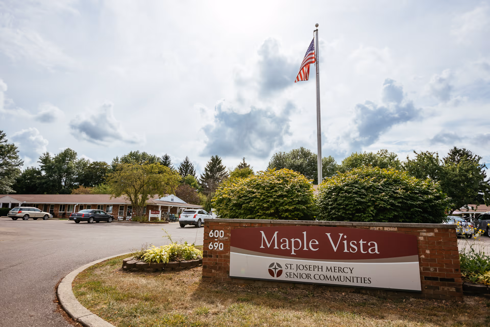 Exterior view of Maple Vista Senior Community with a brick sign, flagpole, and cars.