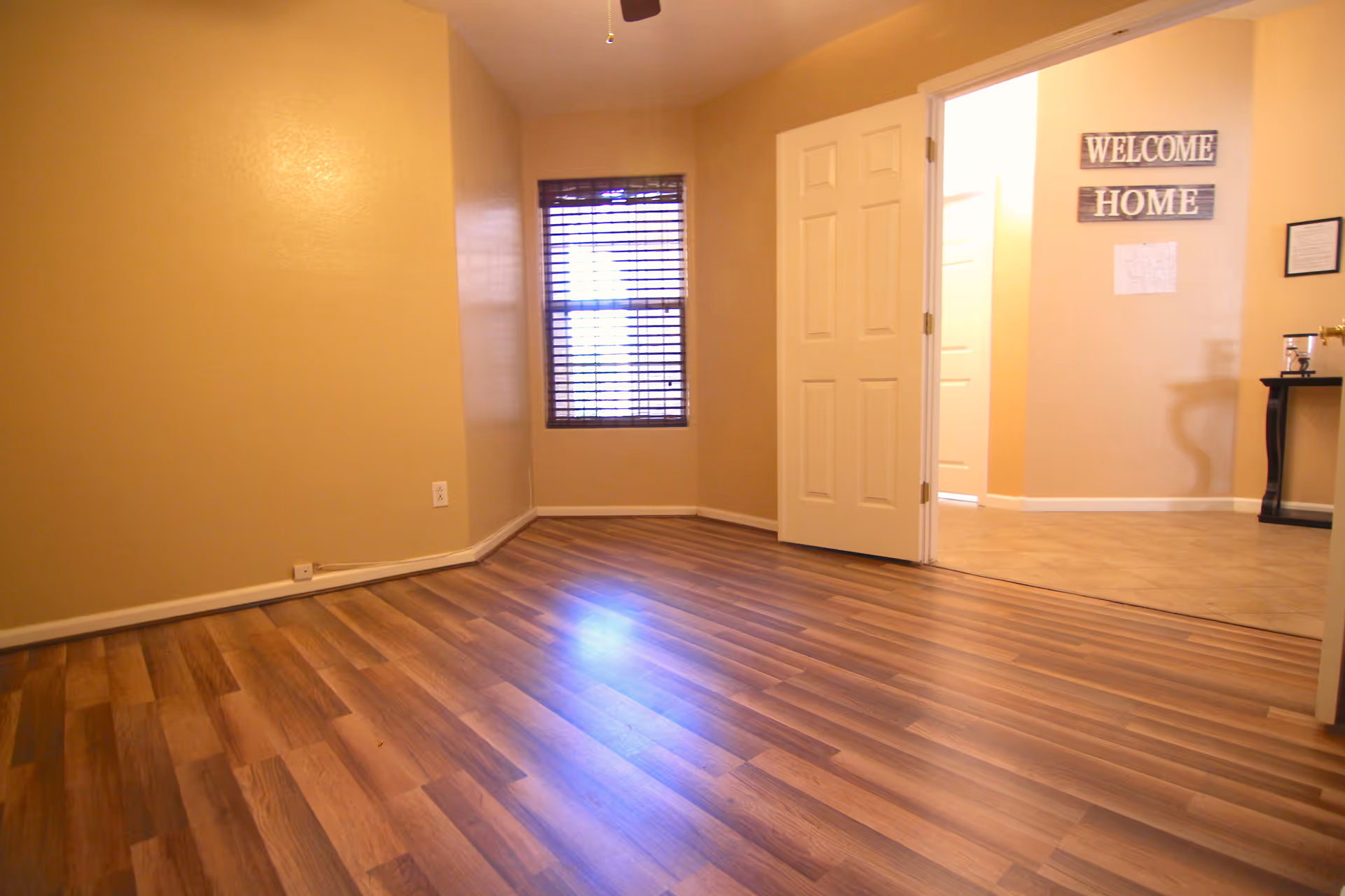 Empty room with wood laminate flooring, beige walls, a window with wooden blinds, and an open door leading to a hallway with a 'WELCOME HOME' sign on the wall.