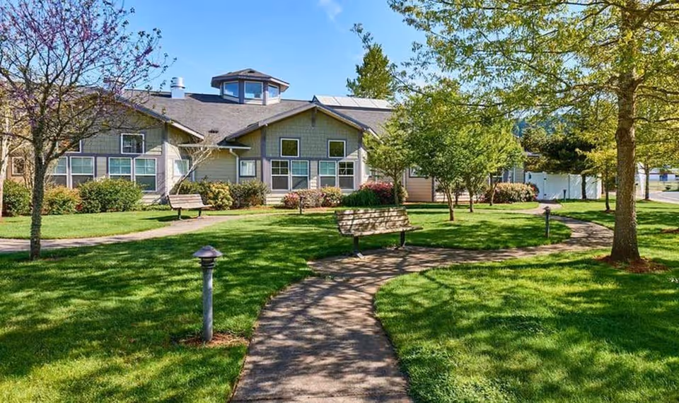 A landscaped courtyard with winding paved paths, benches, trees, and the front of a single-story assisted living building under a blue sky.