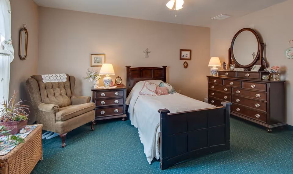 A cozy bedroom with a single wooden bed covered with a white quilt and decorative pillows. To the left of the bed is a wooden nightstand with a lamp, clock, and flower vase. Next to the nightstand is a cushioned armchair with a lace doily on the backrest. On the right side of the room is a large wooden dresser with an oval mirror, a lamp, and decorative items. The room has green carpet flooring and beige walls with framed pictures and a cross hanging above the bed.