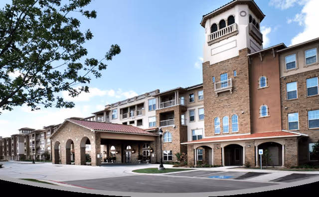 Exterior view of a multi-story senior living facility with a covered entrance, brick and stucco facade, and a tower-like structure. The building is surrounded by a paved driveway and some landscaping, with a tree visible on the left side under a partly cloudy sky.