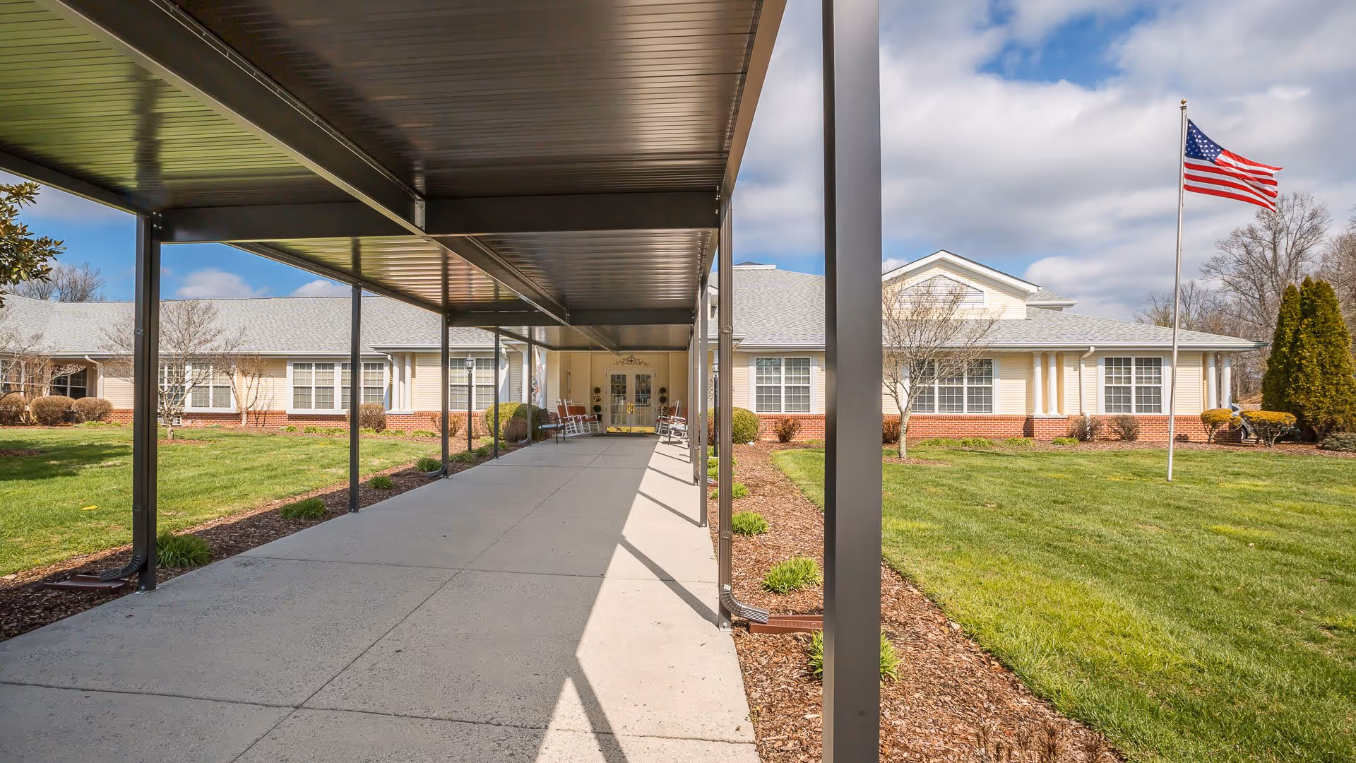 Covered walkway leading to the entrance of a single-story building with beige siding and brick accents, surrounded by green lawns and landscaping. An American flag is flying on a flagpole to the right of the walkway.