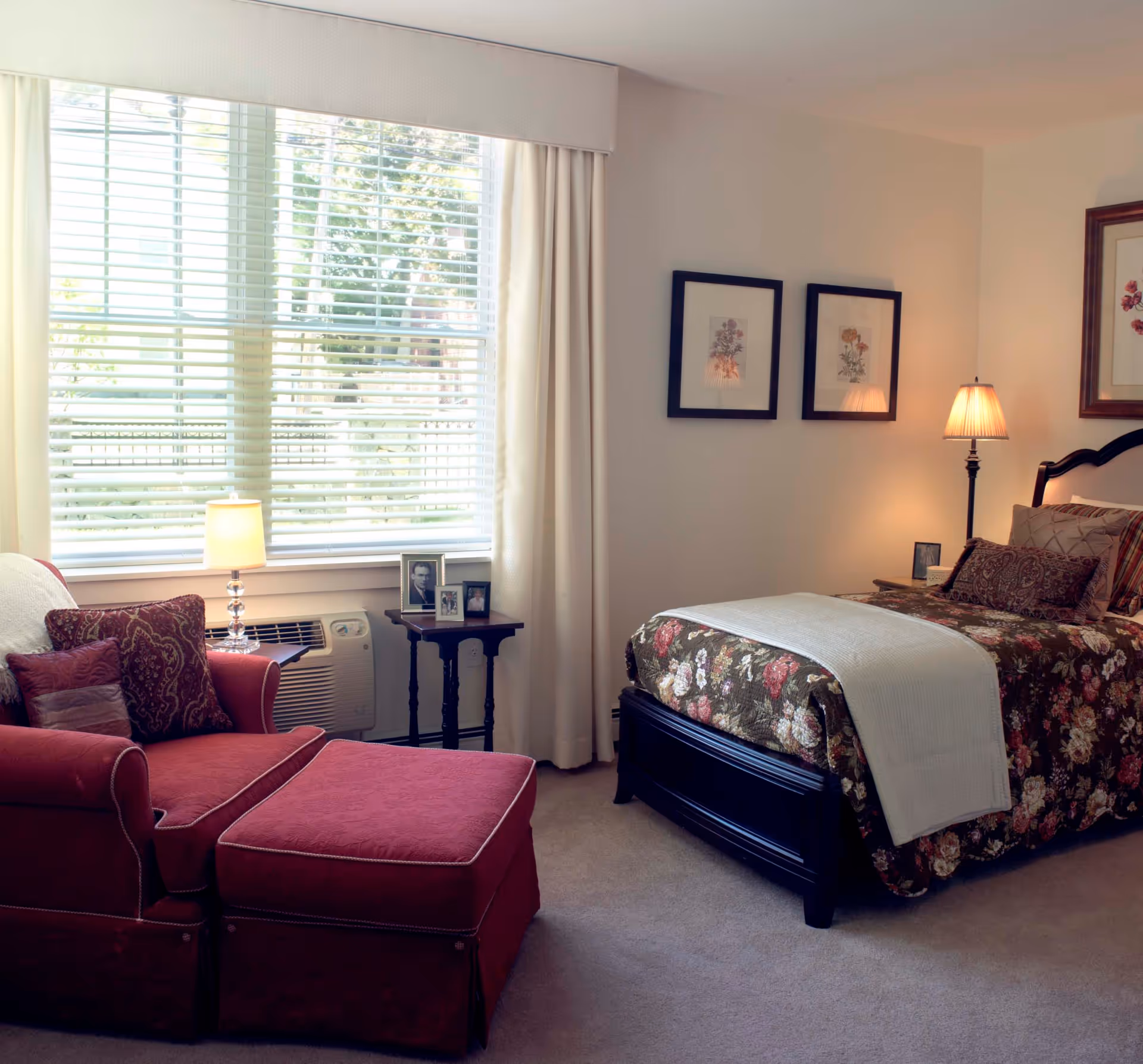 A cozy bedroom with a floral-patterned bedspread on a dark wooden bed frame, a matching armchair with an ottoman in red fabric, a small side table with framed photos, a lamp, and a large window with white blinds and curtains letting in natural light.