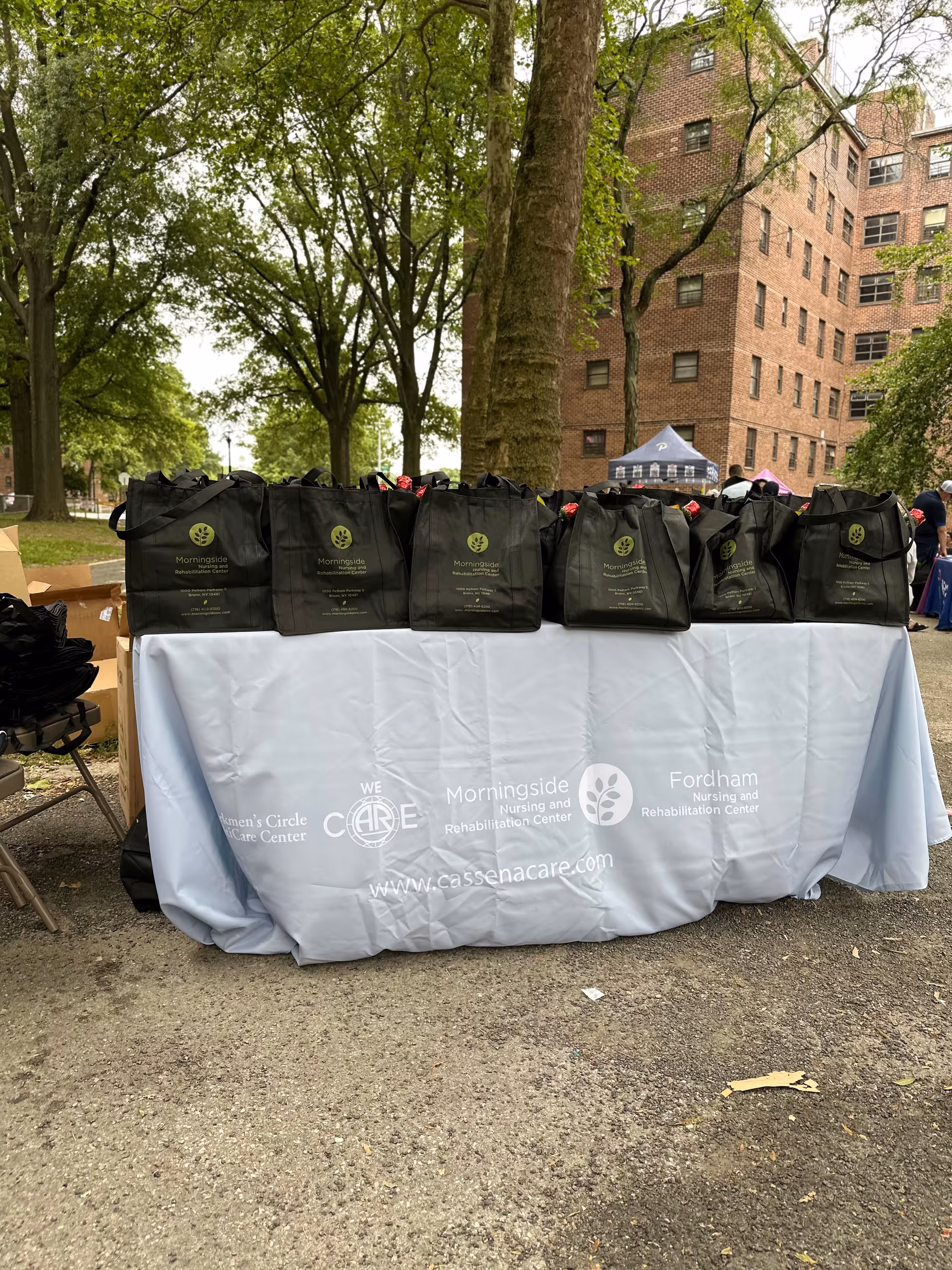 Outdoor event table with black tote bags and a branded cloth near a brick building.