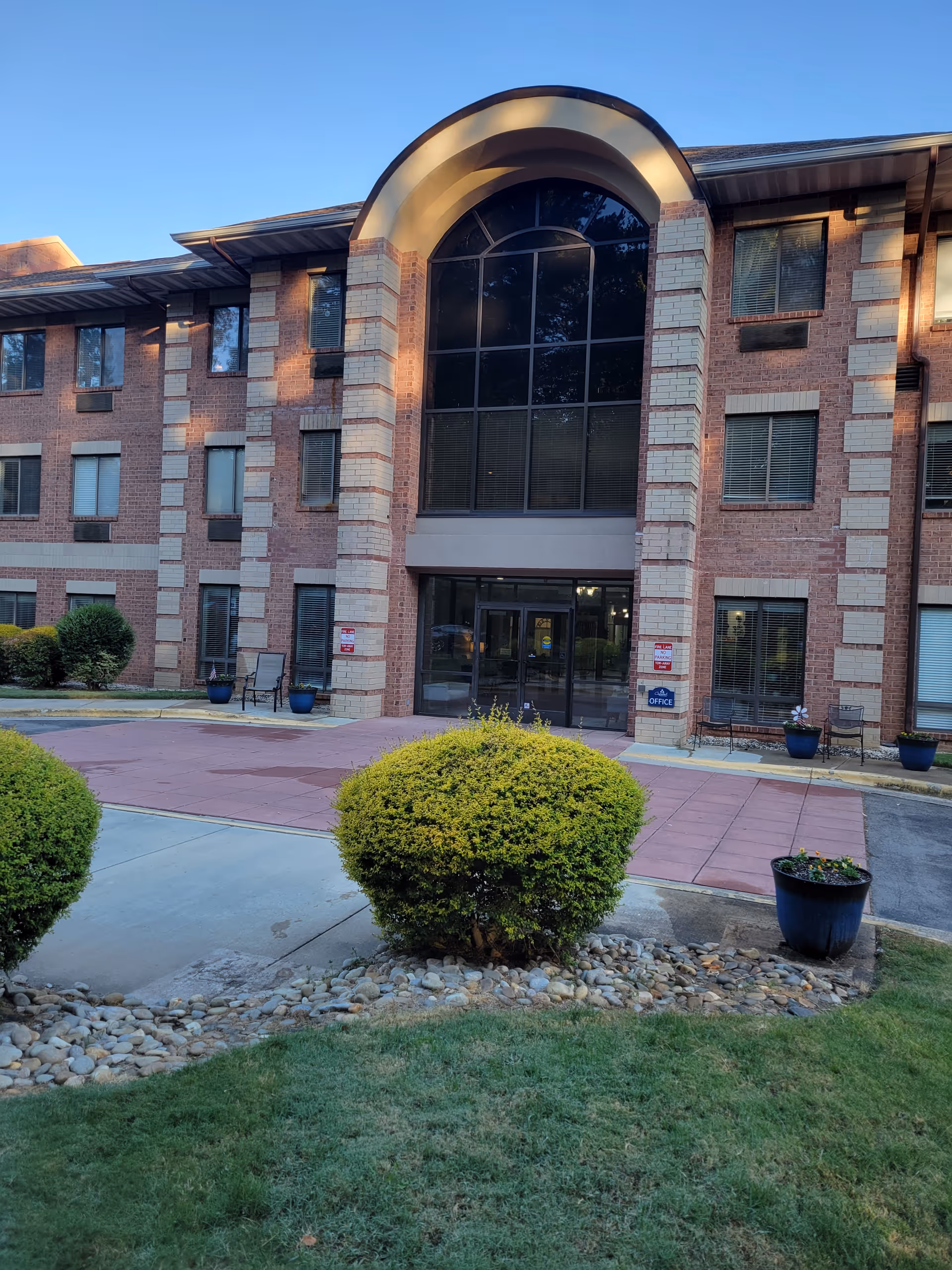 Front exterior view of a three-story brick building with large arched windows above the entrance. There are bushes, potted plants, and a paved walkway leading to the glass double doors. The building has multiple windows with blinds and a sign near the entrance indicating the office.