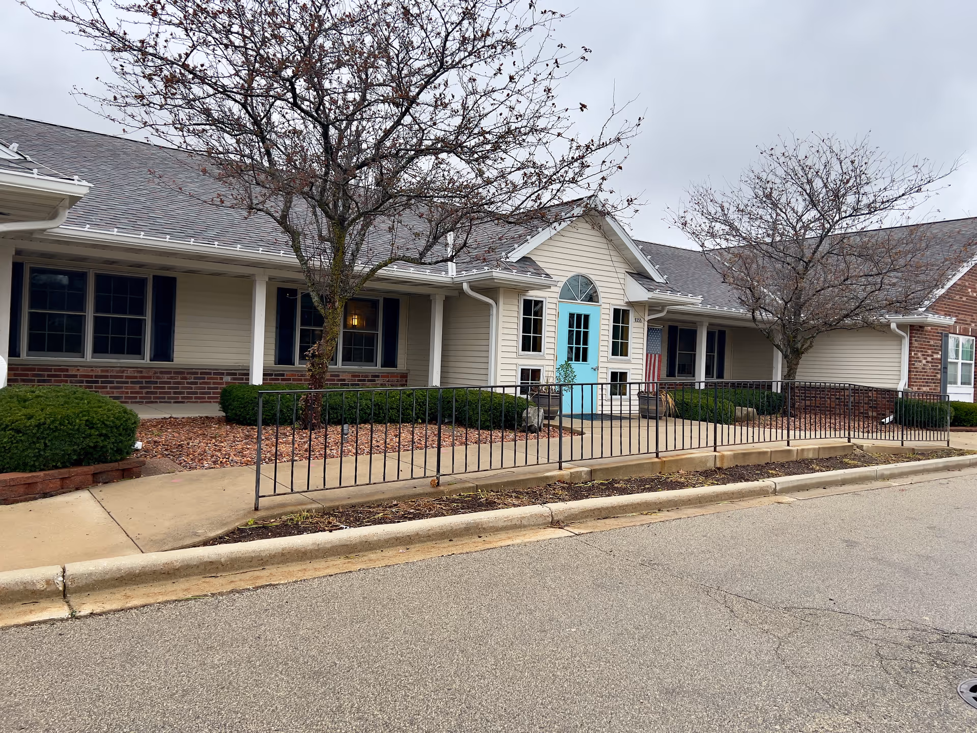 Exterior view of a single-story building with beige siding and brick accents, featuring a bright turquoise front door with an arched window above it. There are leafless trees and neatly trimmed bushes in front of the building, along with a black metal railing along the sidewalk leading to the entrance. The sky is overcast.