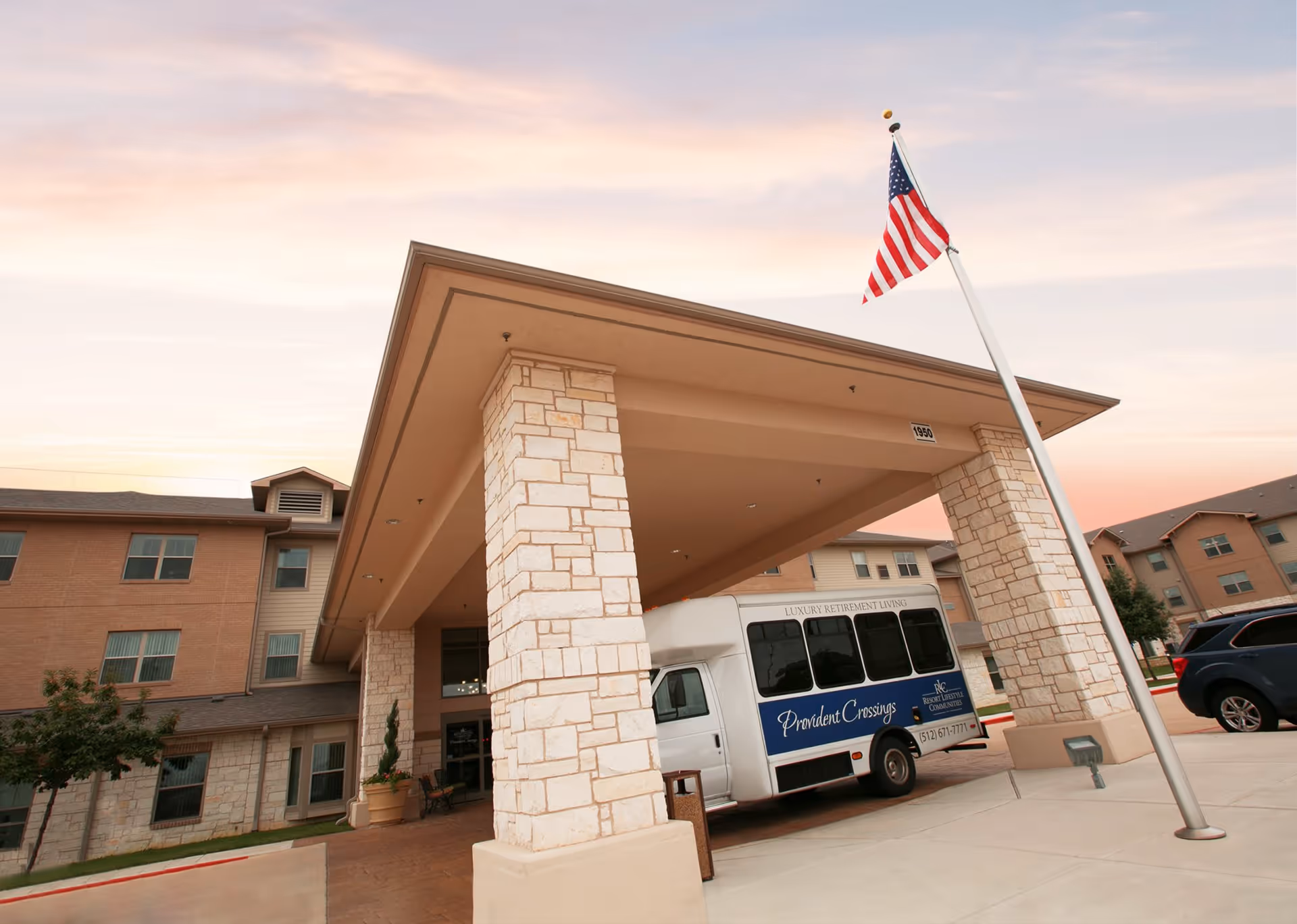 Entrance of Provident Crossings Retirement Resort with a covered driveway supported by stone pillars, an American flag on a tall flagpole, a shuttle bus parked under the canopy, and a multi-story building in the background during sunset.