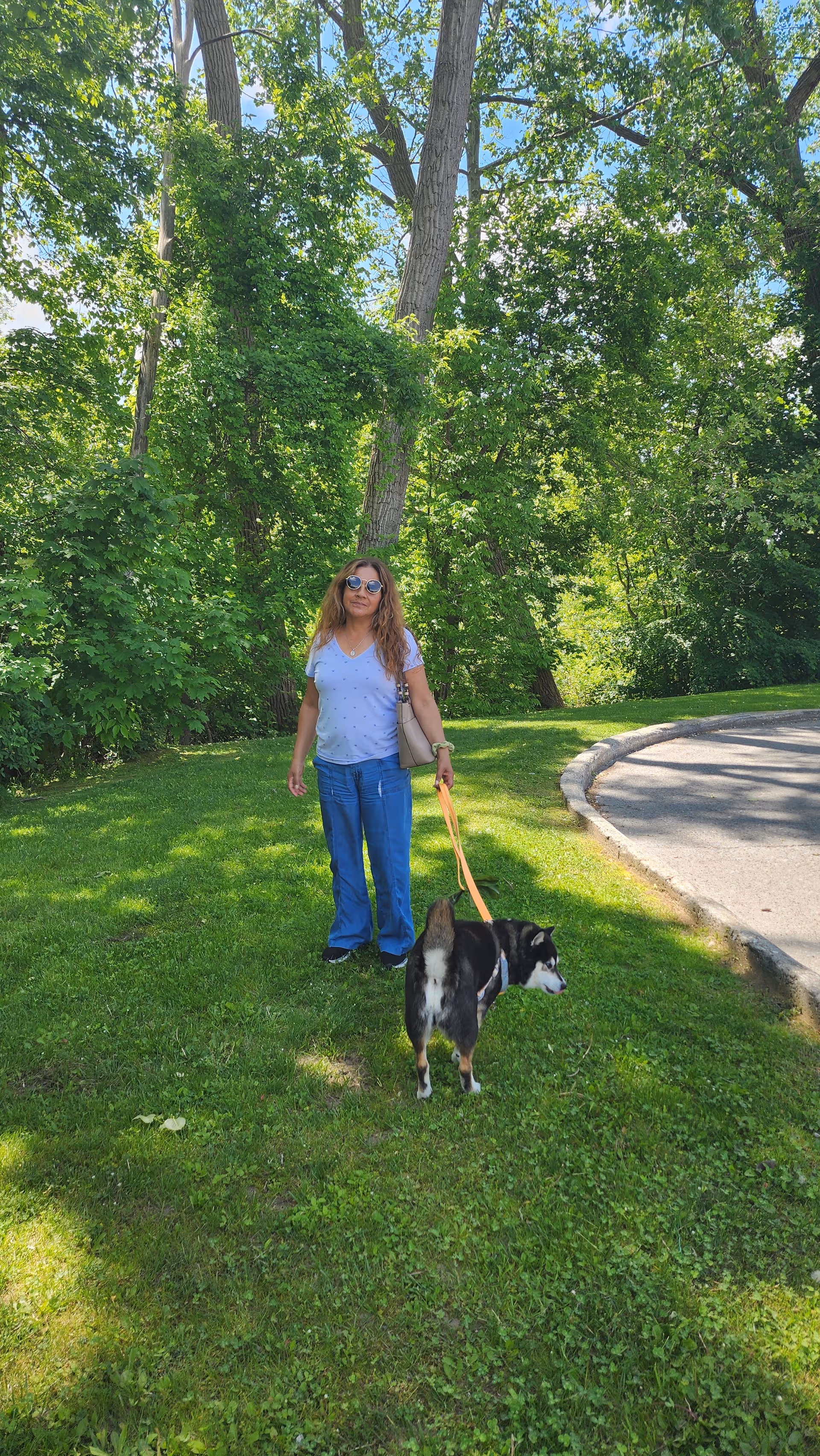 A woman stands on a grassy outdoor area with a leashed dog beside a path.