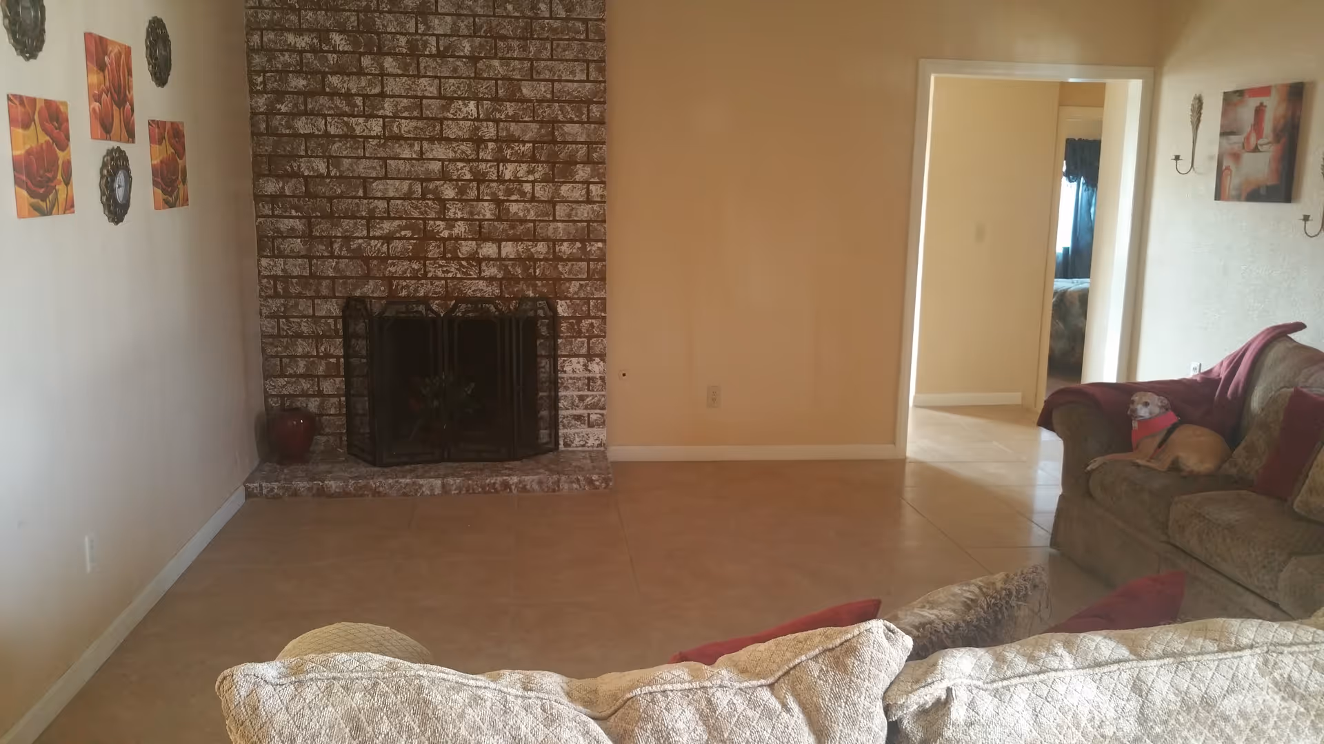 A living room with beige tiled floor and light-colored walls. There is a brick fireplace with a black metal screen in the center of the far wall. On the left wall, there are three floral paintings and three decorative round wall hangings. To the right, there is a doorway leading to another room with a bed visible. A brown couch with red cushions and a red throw blanket is on the right side, with a small dog wearing a pink harness sitting on it. In the foreground, the back of another couch with beige cushions is visible.
