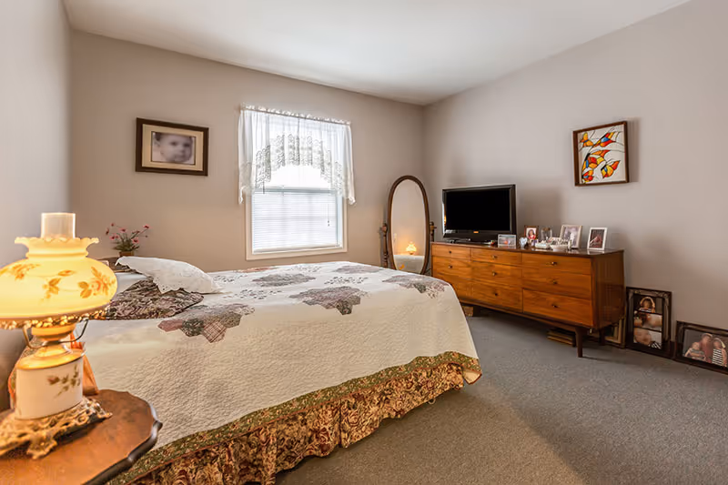 A cozy bedroom with a large bed, dresser, TV, and window in soft lighting.
