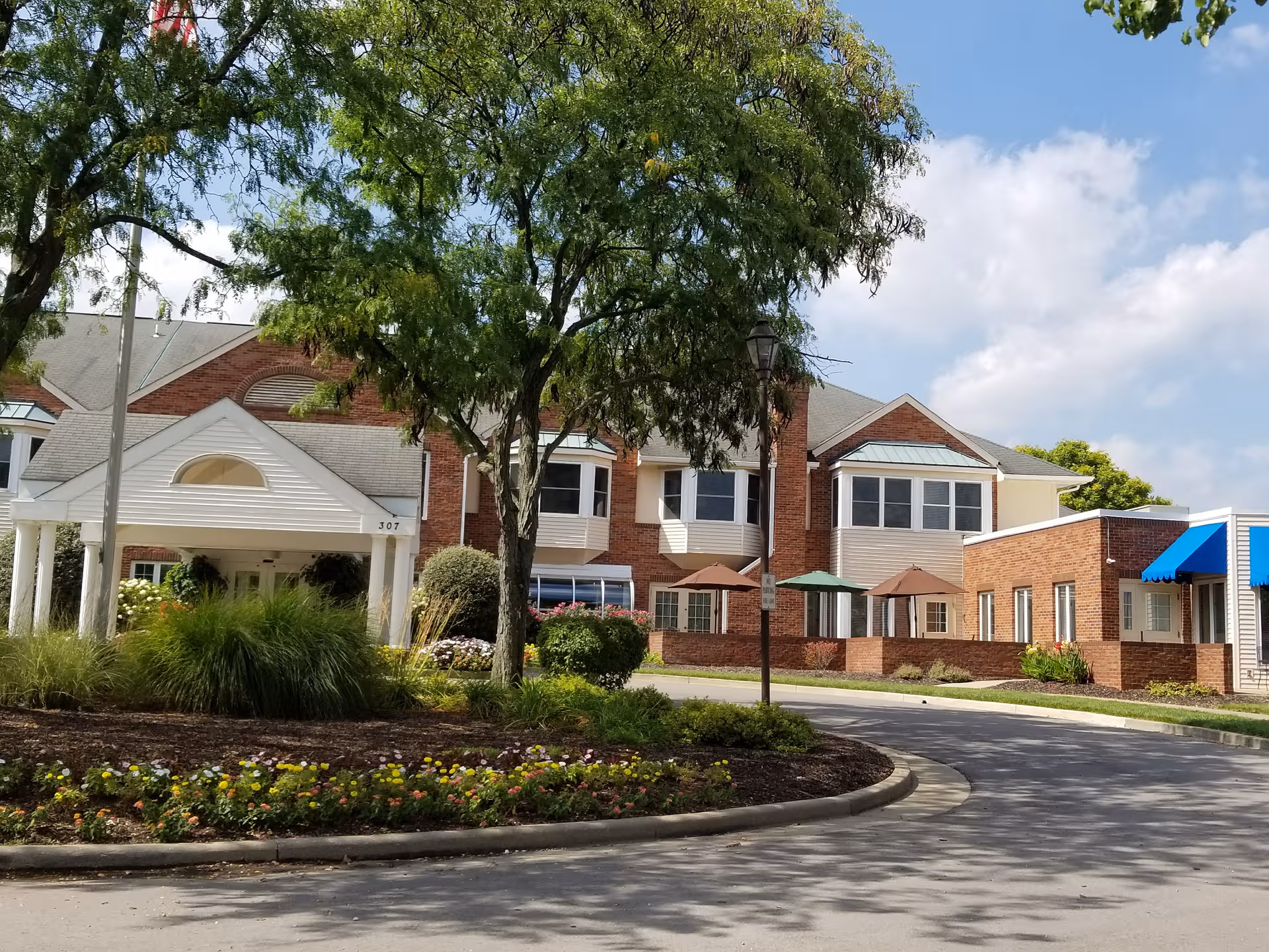 Brick senior living building with landscaped entrance, curved driveway, trees, and outdoor umbrellas.
