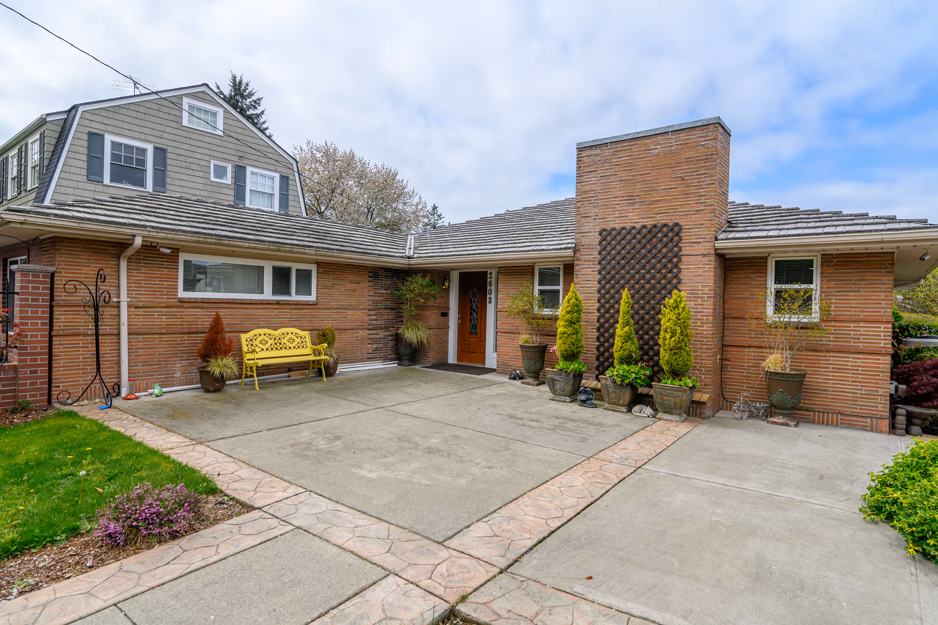 Front exterior of a brick assisted living facility with a driveway, bench, and potted plants.