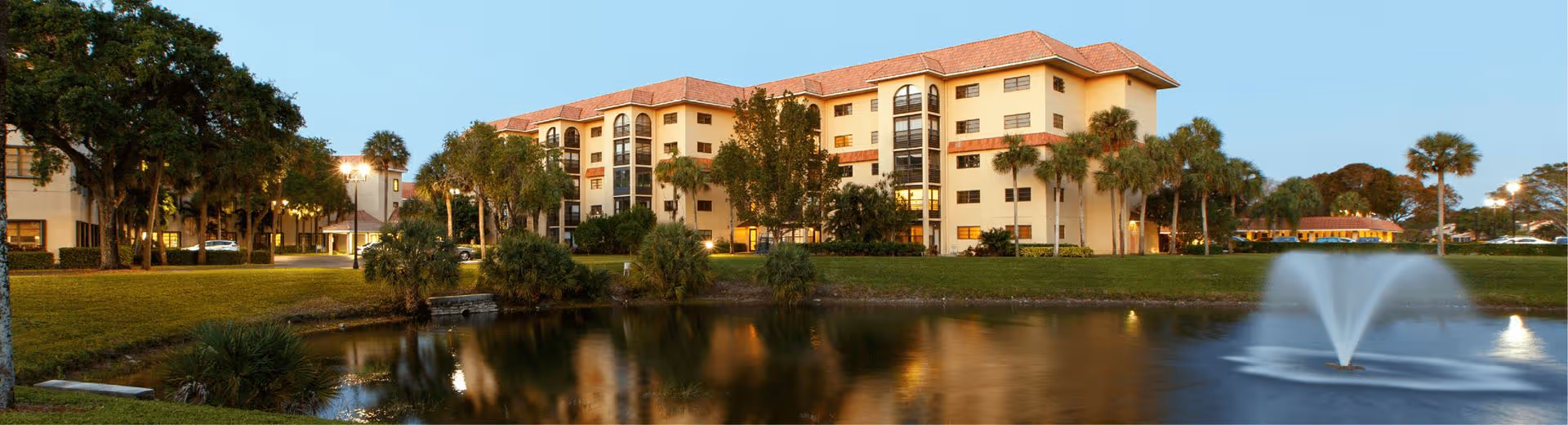 Multi-story senior living building beside a pond with a fountain and palm trees at dusk.