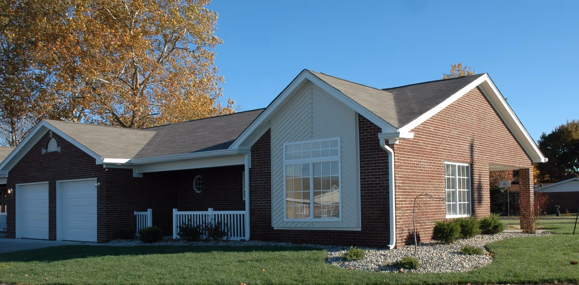 Exterior view of a single-story brick building with a gable roof, white trim, and large windows. The building has a two-car garage with white doors, a small porch with white railing, and landscaping with grass and shrubs. Trees with autumn leaves are visible in the background under a clear blue sky.