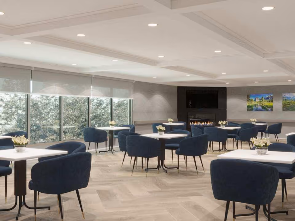 Bright communal dining room with round tables, blue upholstered chairs, large windows, and a fireplace.