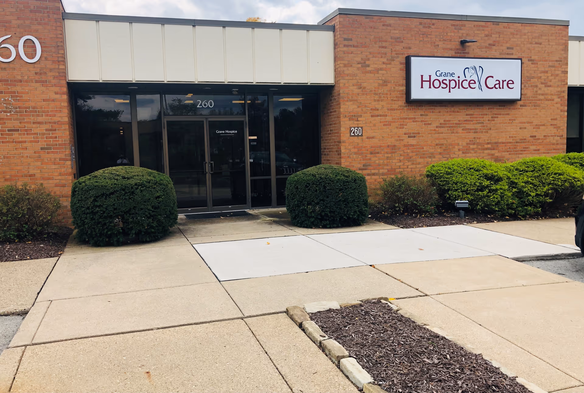 Brick building entrance with Grane Hospice Care sign, glass doors, and trimmed shrubs.