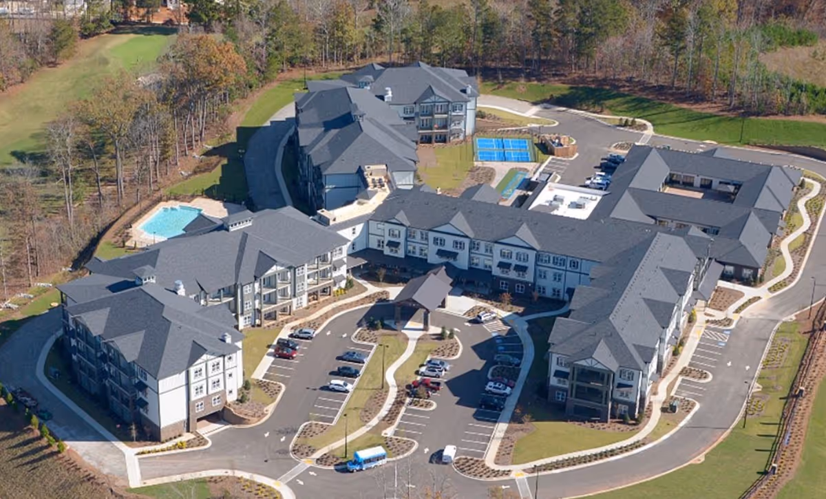 Aerial view of a senior living campus with multiple connected buildings, parking lots, a swimming pool and wooded surroundings.