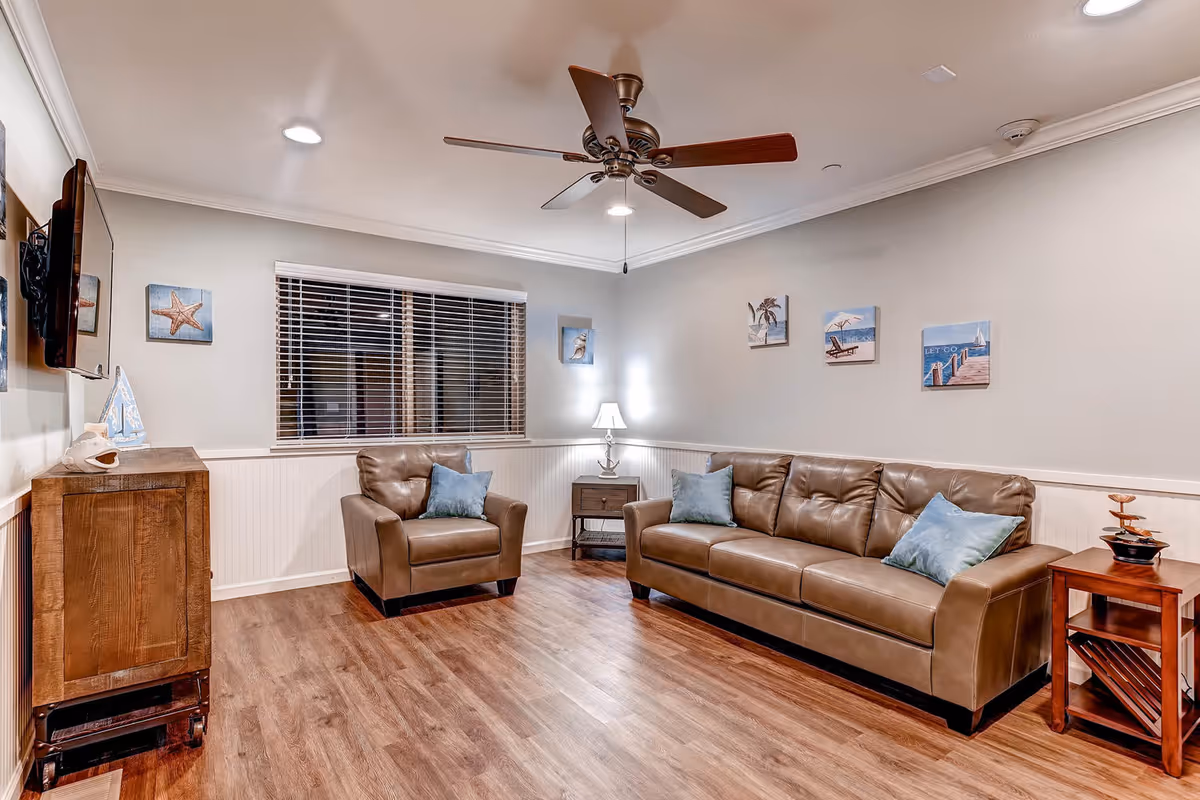 Cozy senior living room with leather sofa set, ceiling fan, and beach-themed wall art.