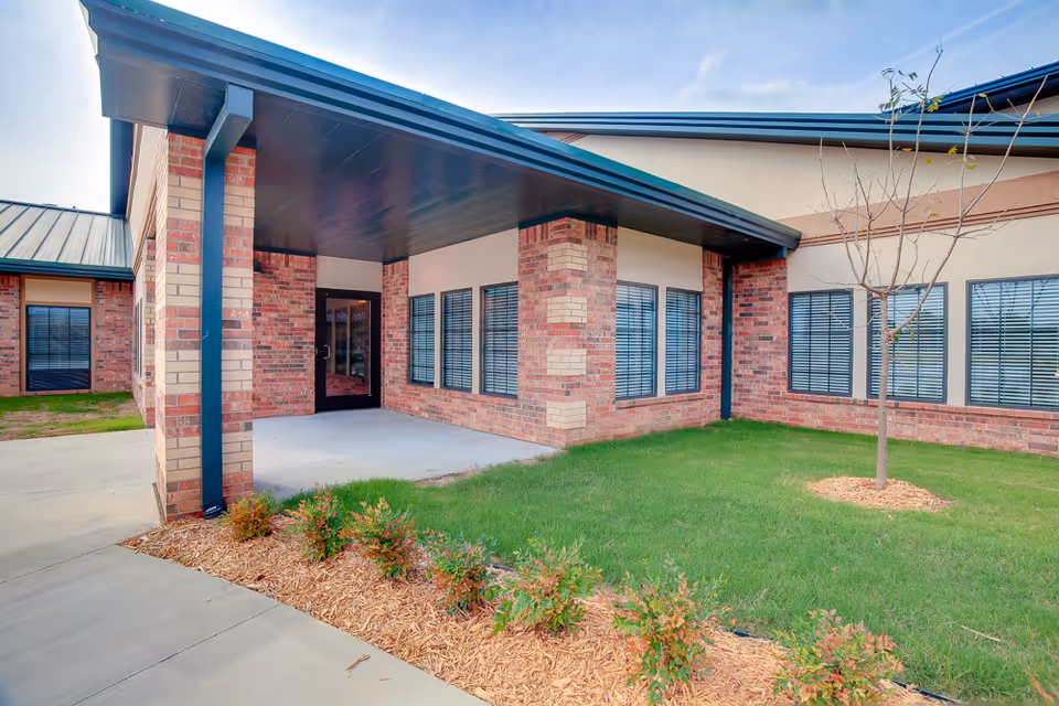 Exterior view of a brick building with a covered entrance, several windows with blinds, a small tree, and landscaped greenery with mulch along a concrete walkway under a partly cloudy sky.